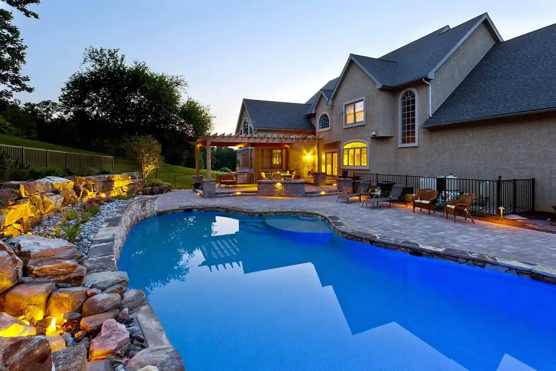 Backyard pool beside a stone patio and house at dusk