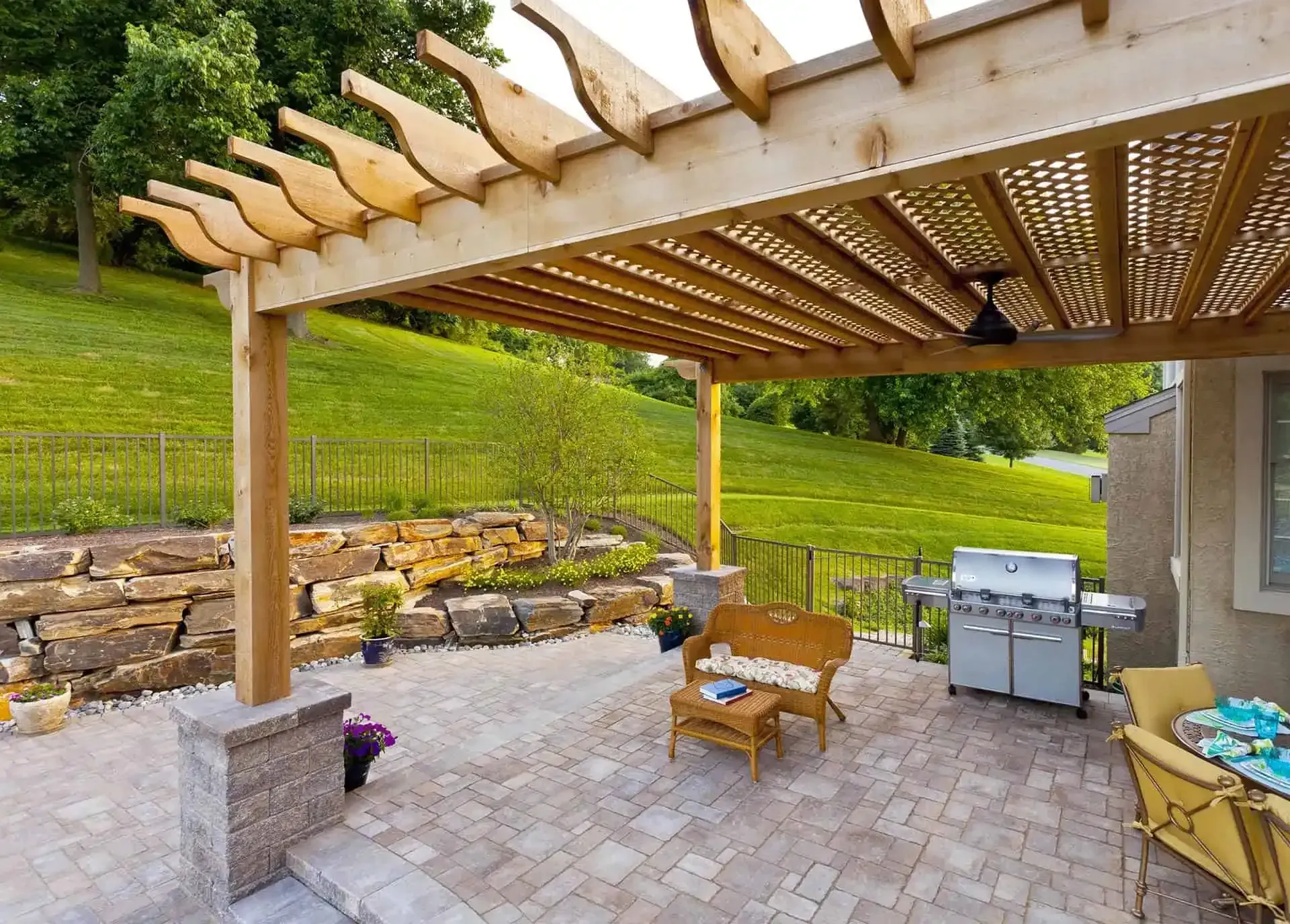 Patio with wooden pergola, wicker chairs, grill, and stone pavers beside a grassy hillside