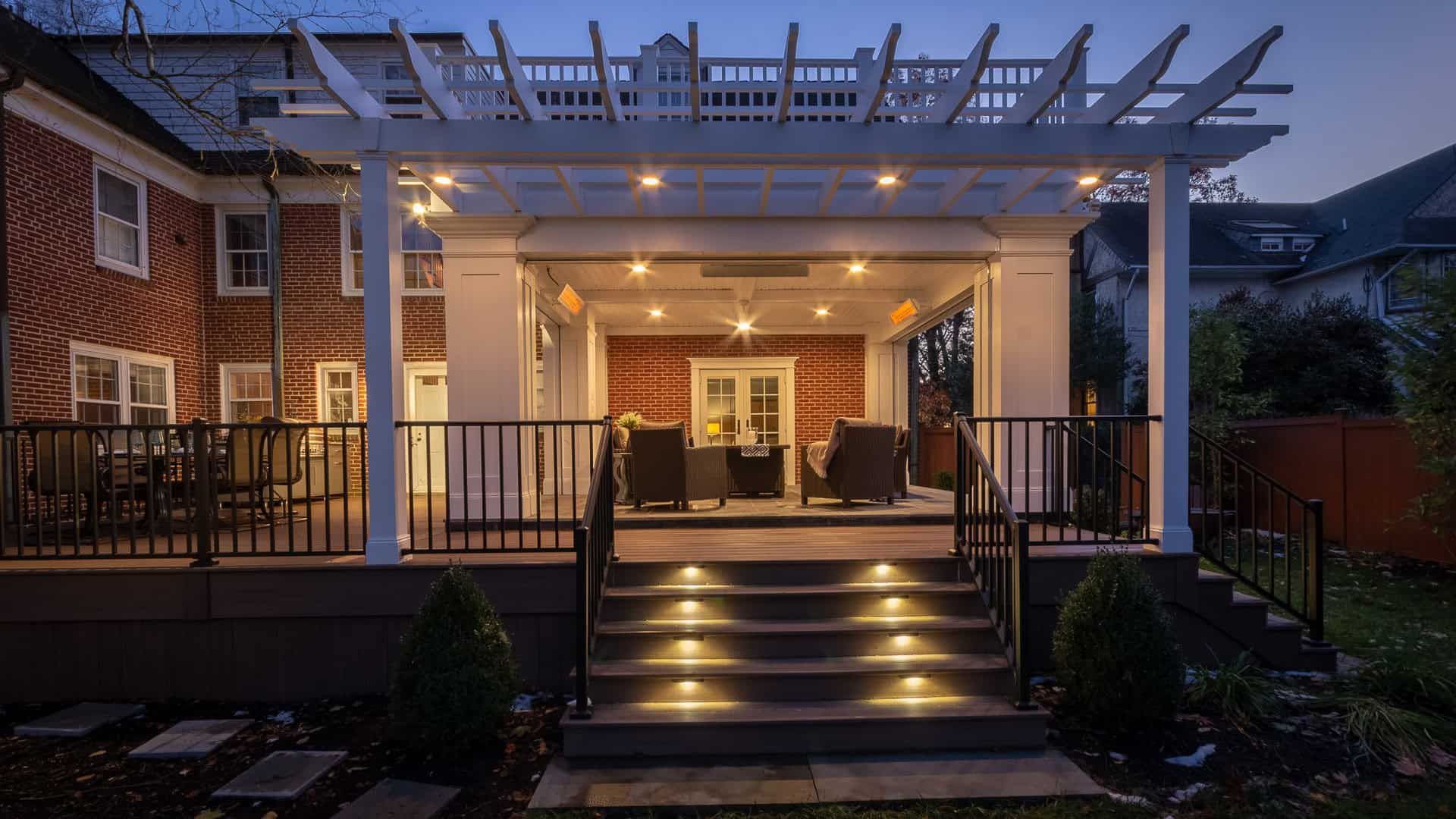 Brick house porch at dusk with lit steps, black railing, and pergola overhead