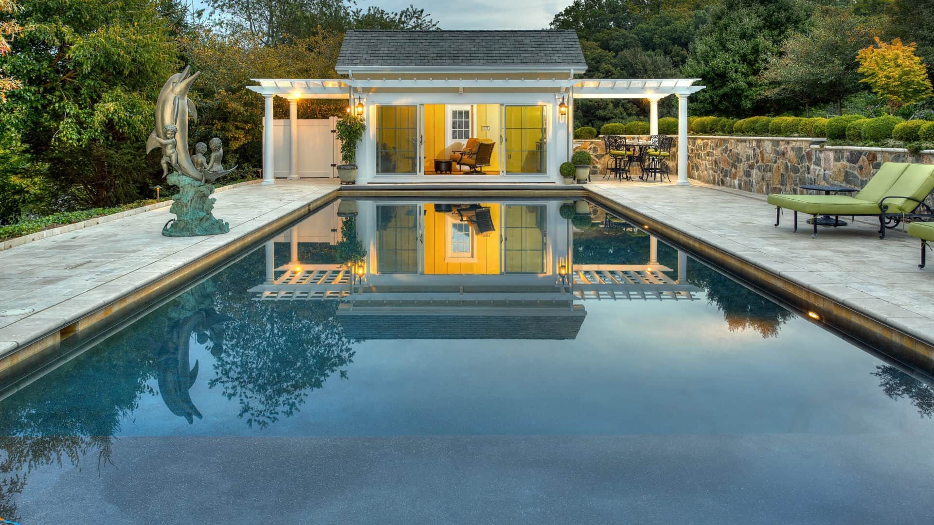 Modern backyard pool with a pool house, stone patio, and lounge chairs at dusk