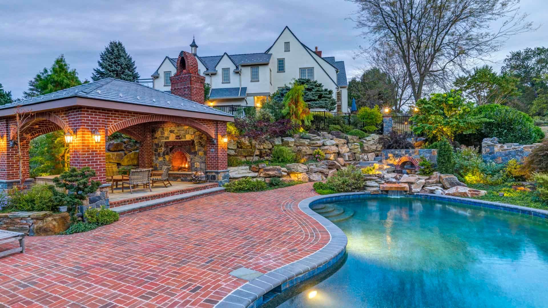 Stone patio and lit gazebo beside a curved backyard pool, with a large house and landscaped garden at dusk
