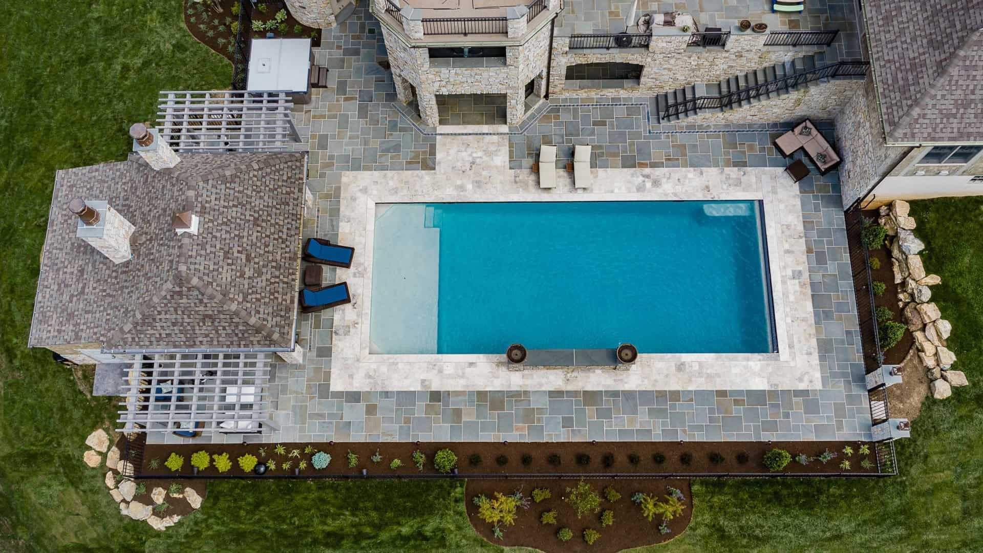 Aerial view of a backyard with a rectangular blue swimming pool, patio seating, and stone walkways