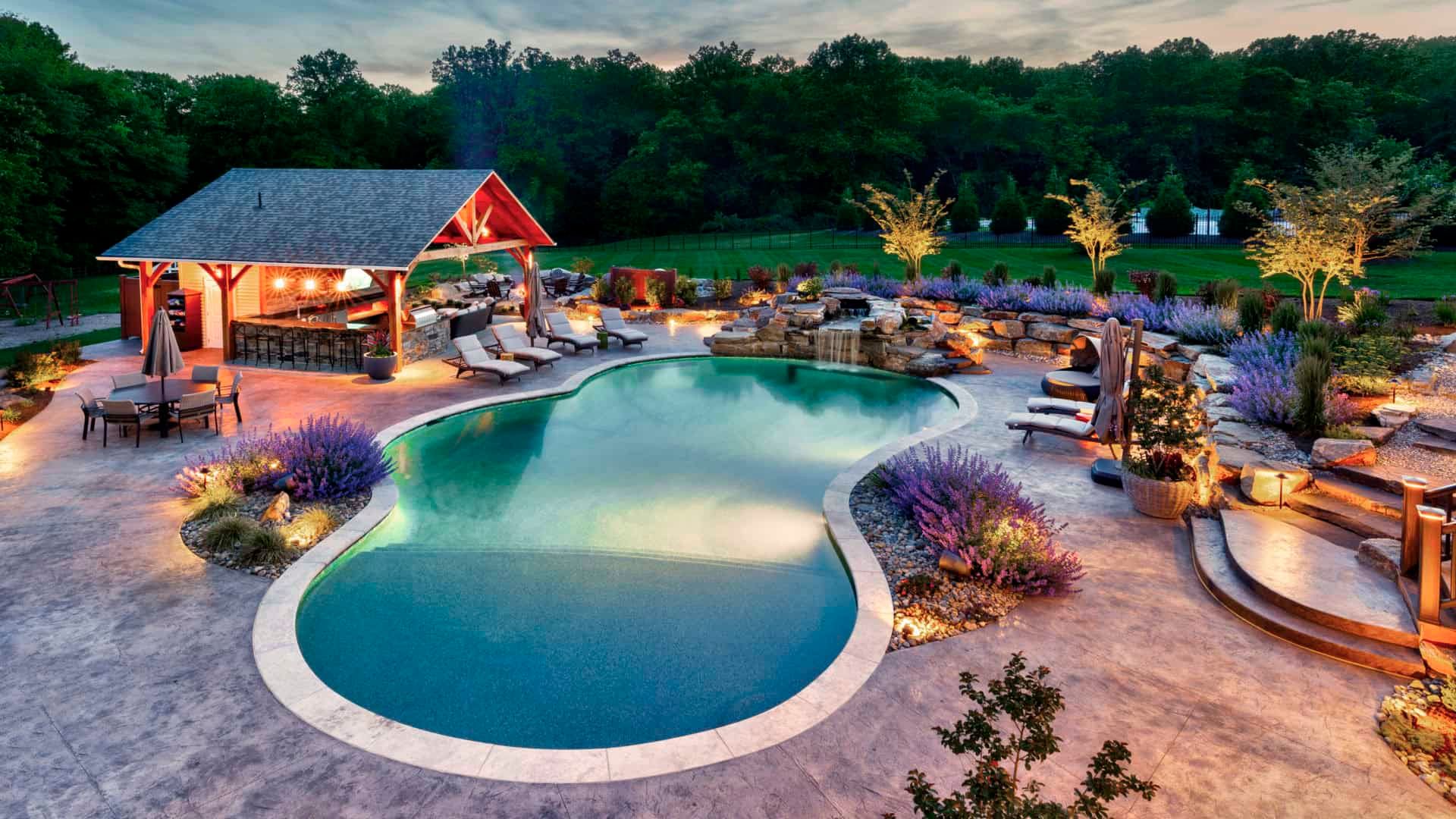Luxury backyard pool with a glowing waterfall, pavilion, and landscaped patio at dusk