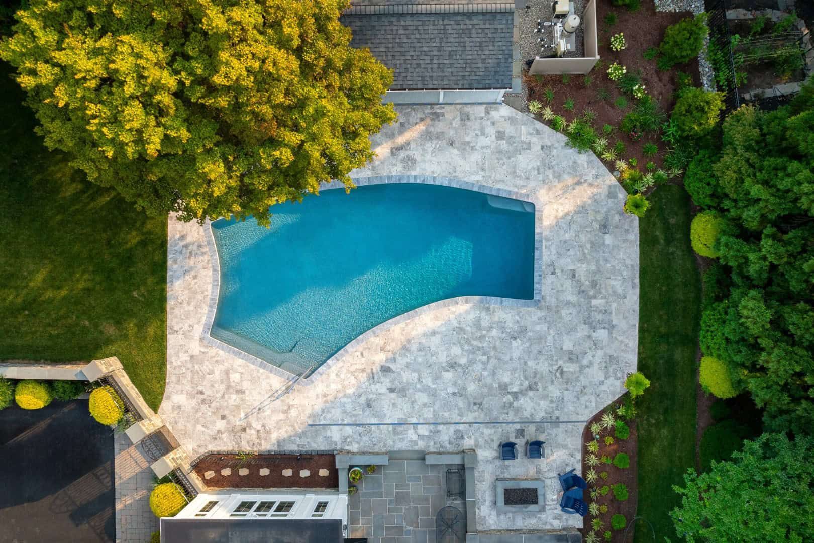 Aerial view of a backyard pool with light stone deck, surrounded by trees and greenery.
