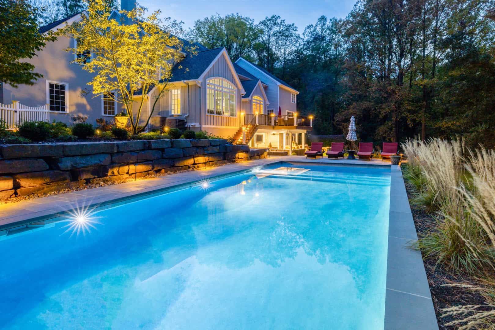 Lit backyard pool beside a modern house at dusk, with warm lights and lounge chairs.