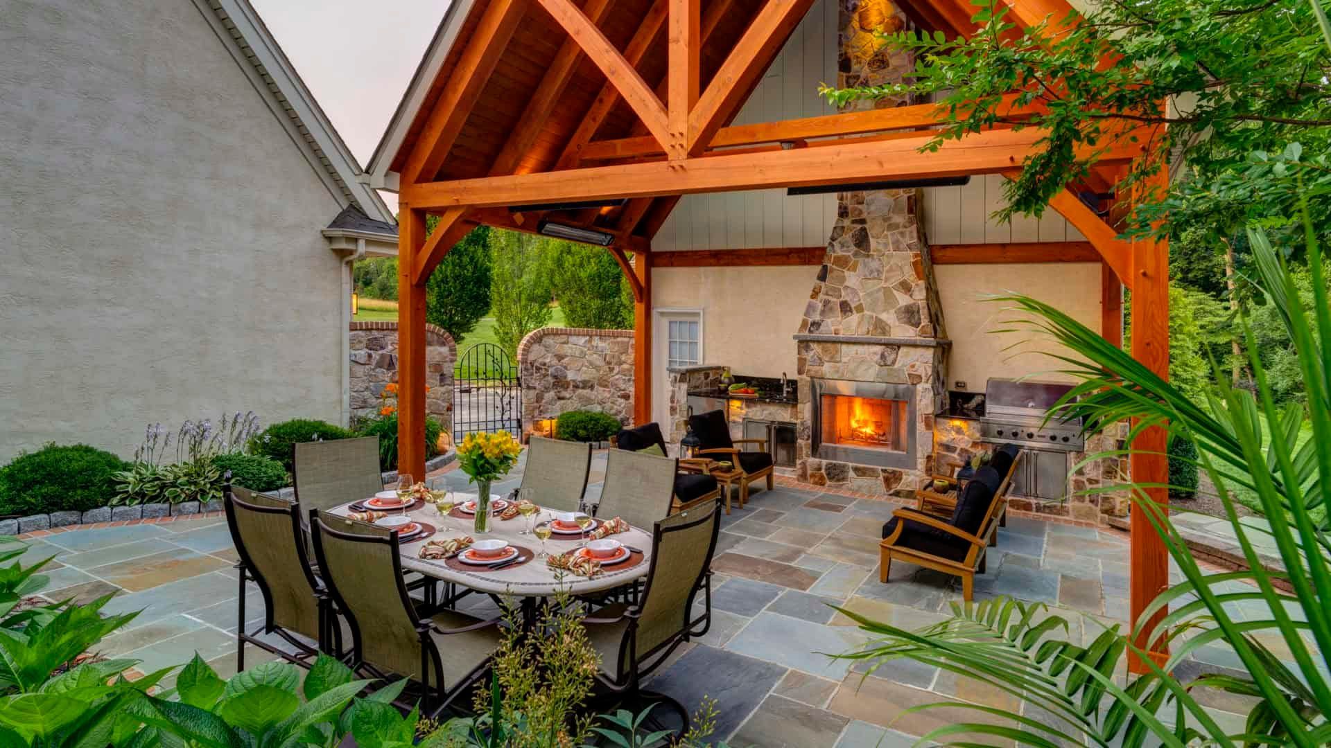 Outdoor stone patio with dining table, chairs, and a lit fireplace under a wooden pergola