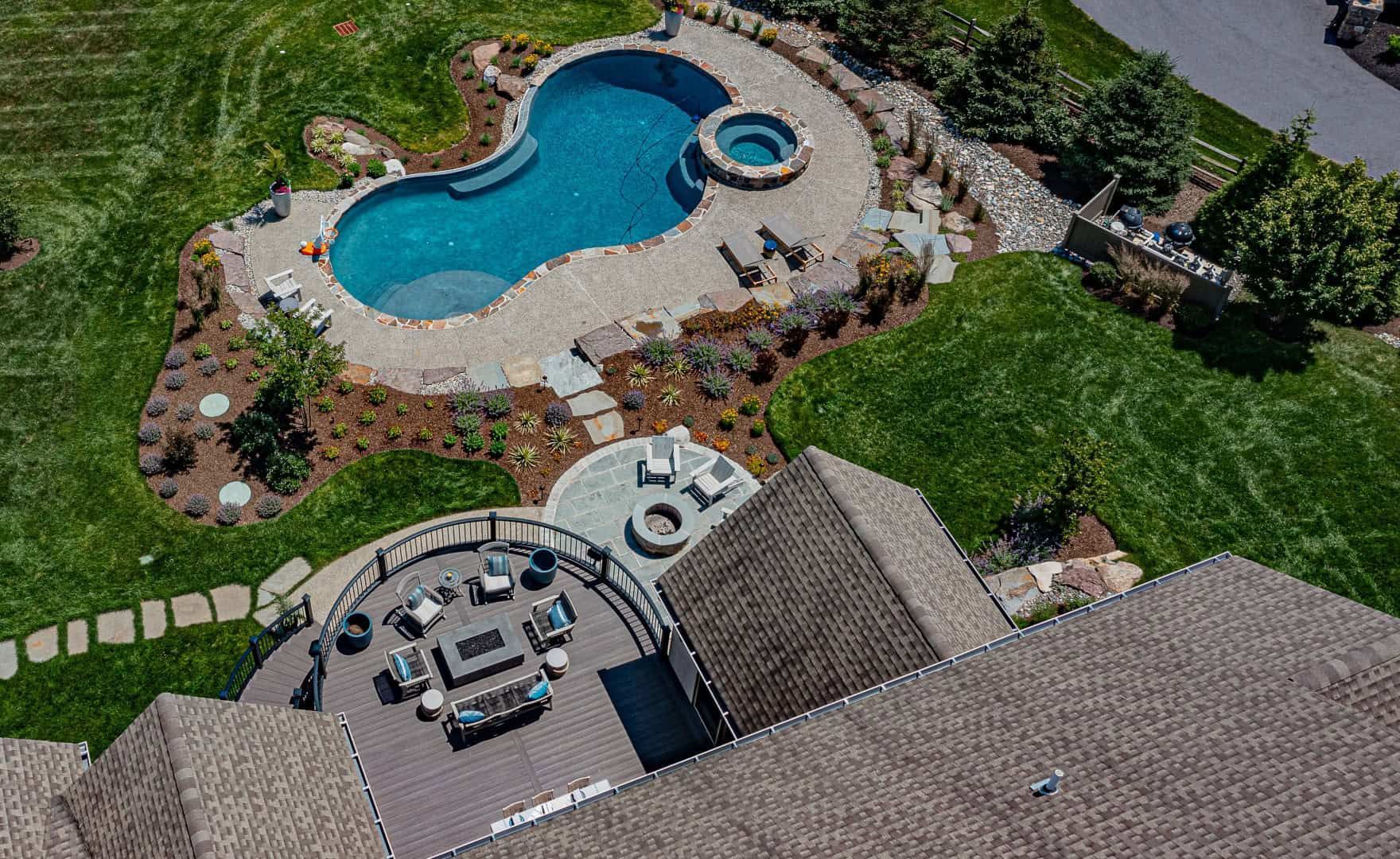 Aerial view of a backyard pool with hot tub, stone patio, and lounge chairs surrounded by green lawn.