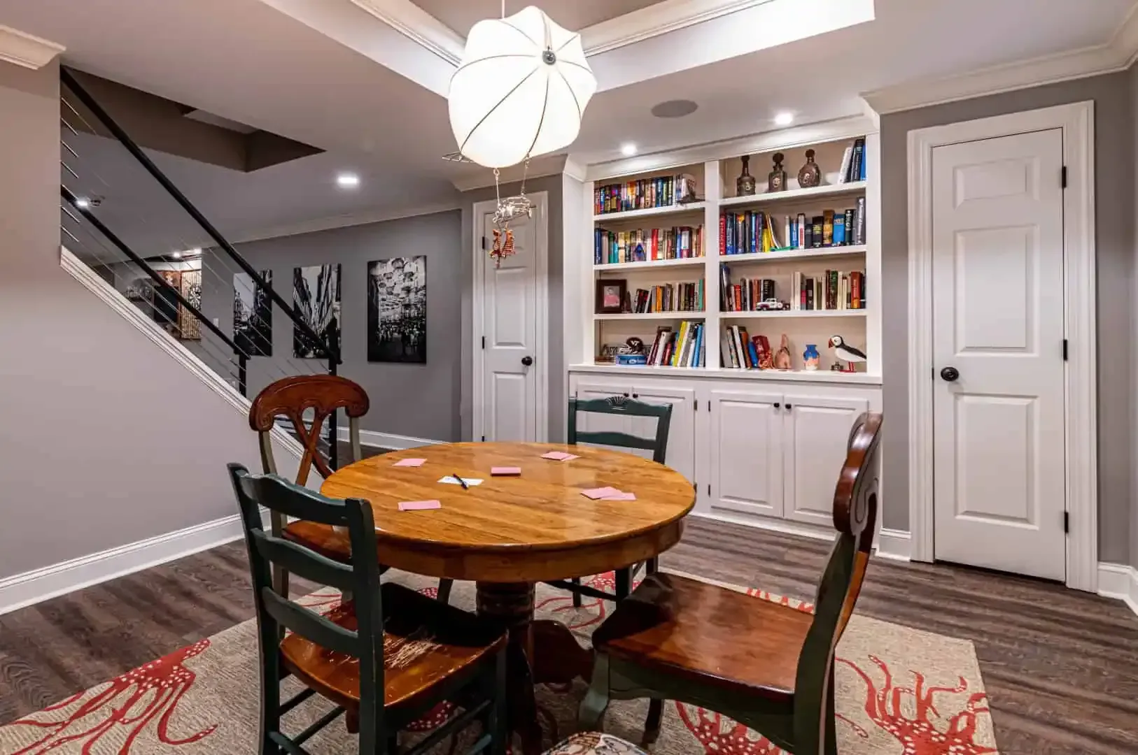 Dining room with round wooden table, chairs, built-in bookshelf, and staircase in the background