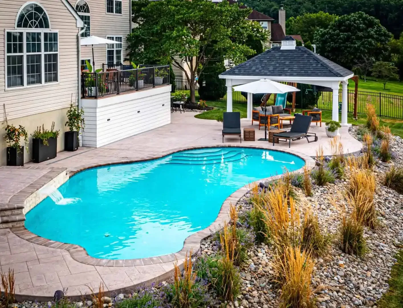 Backyard pool with curved stone deck, patio seating, white umbrella, and landscaped garden beside a house