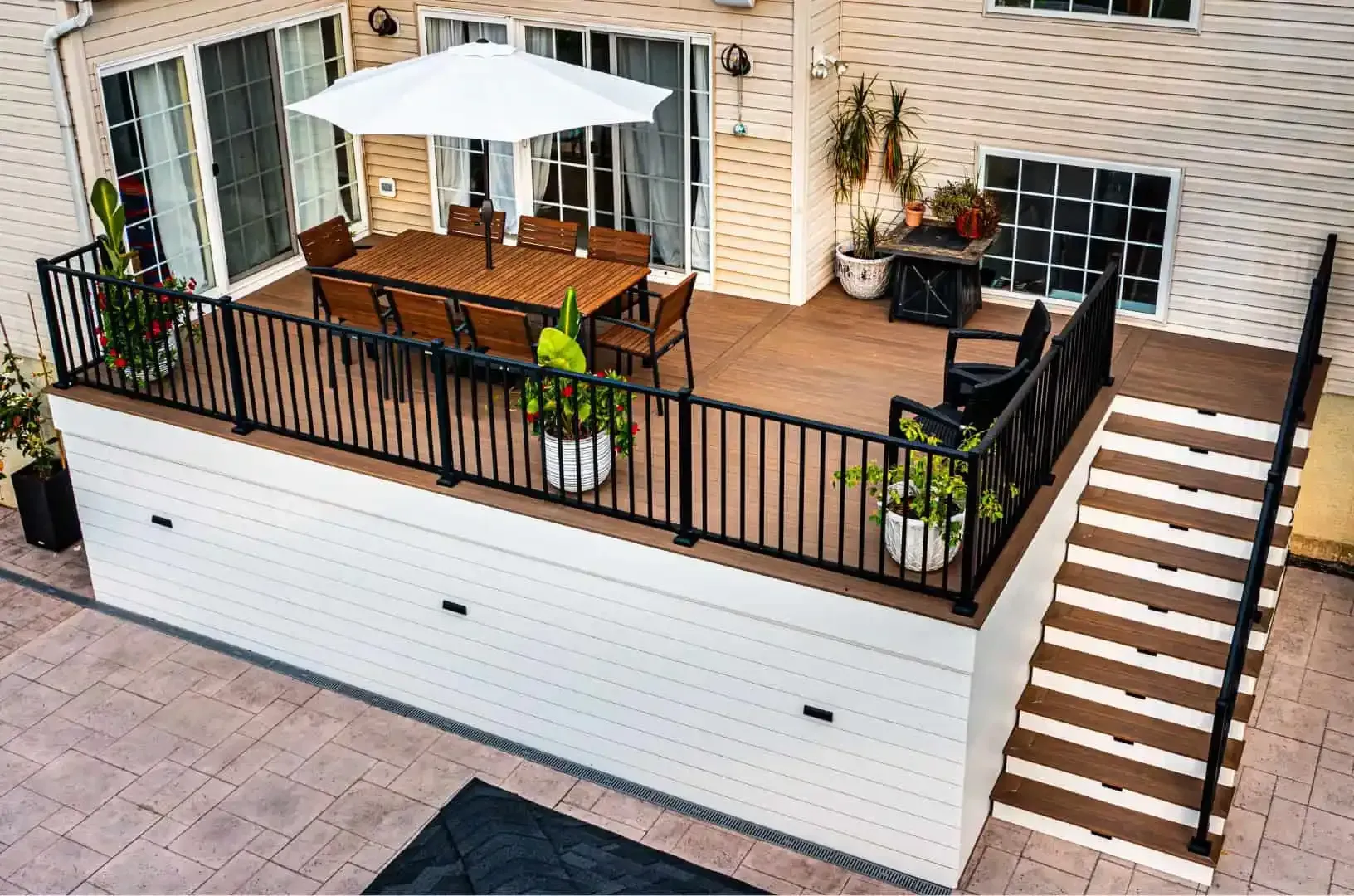 Outdoor patio deck with white umbrella, table, chairs, potted plants, and stairs leading down to a driveway
