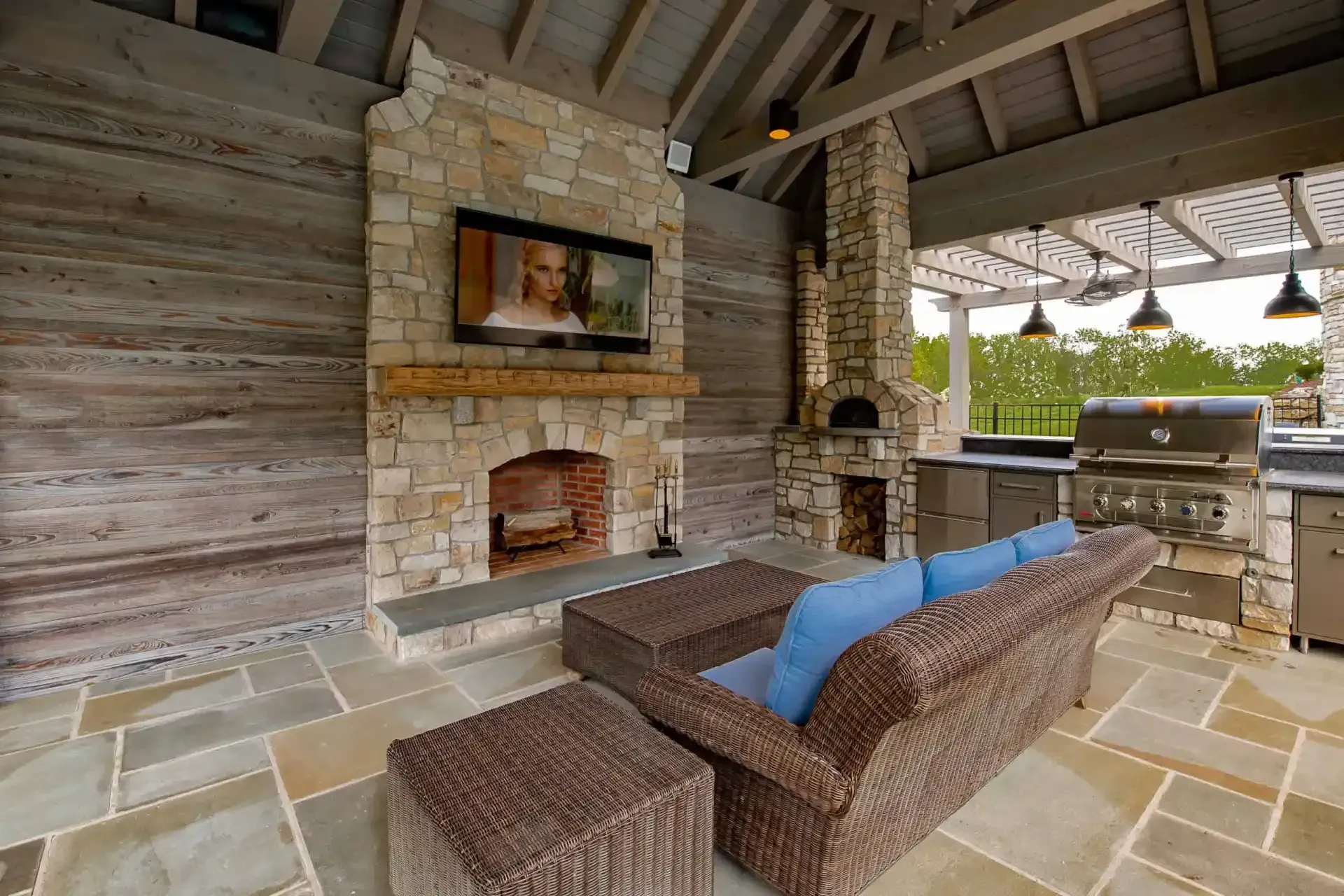 Covered outdoor patio with stone fireplace, wicker sofa, and grill under a wooden roof