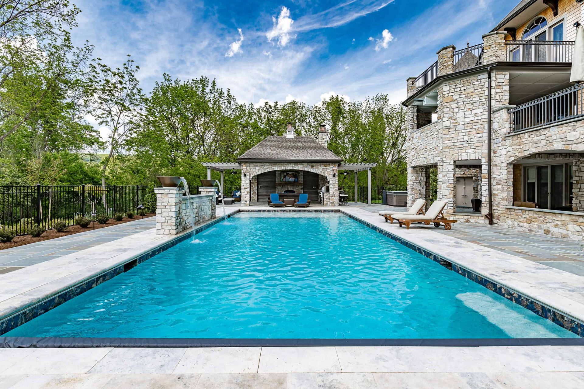 Luxury backyard with blue swimming pool, stone patio, and house exterior under a partly cloudy sky