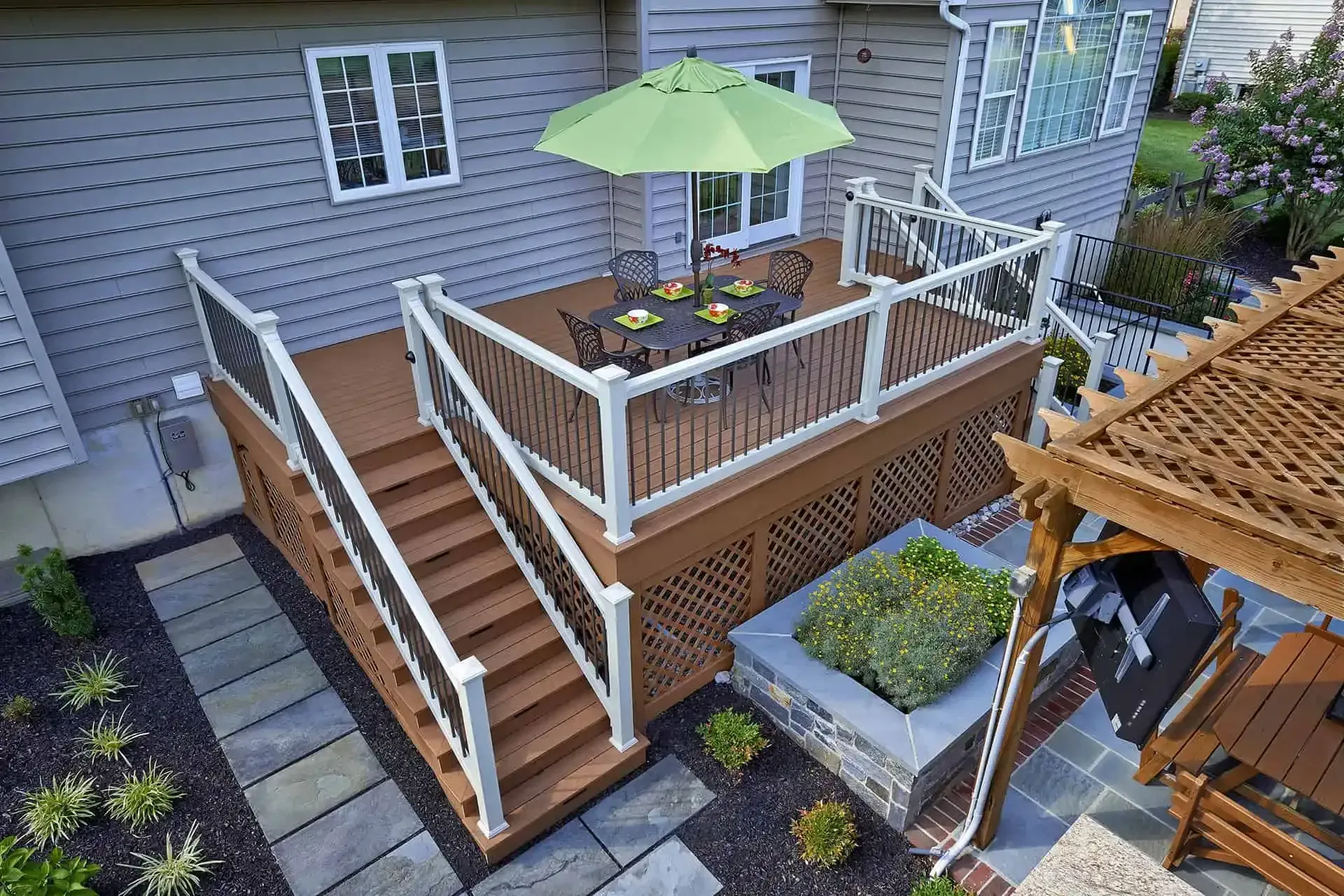 Wooden deck with white railing, outdoor table and green umbrella beside a house and garden steps