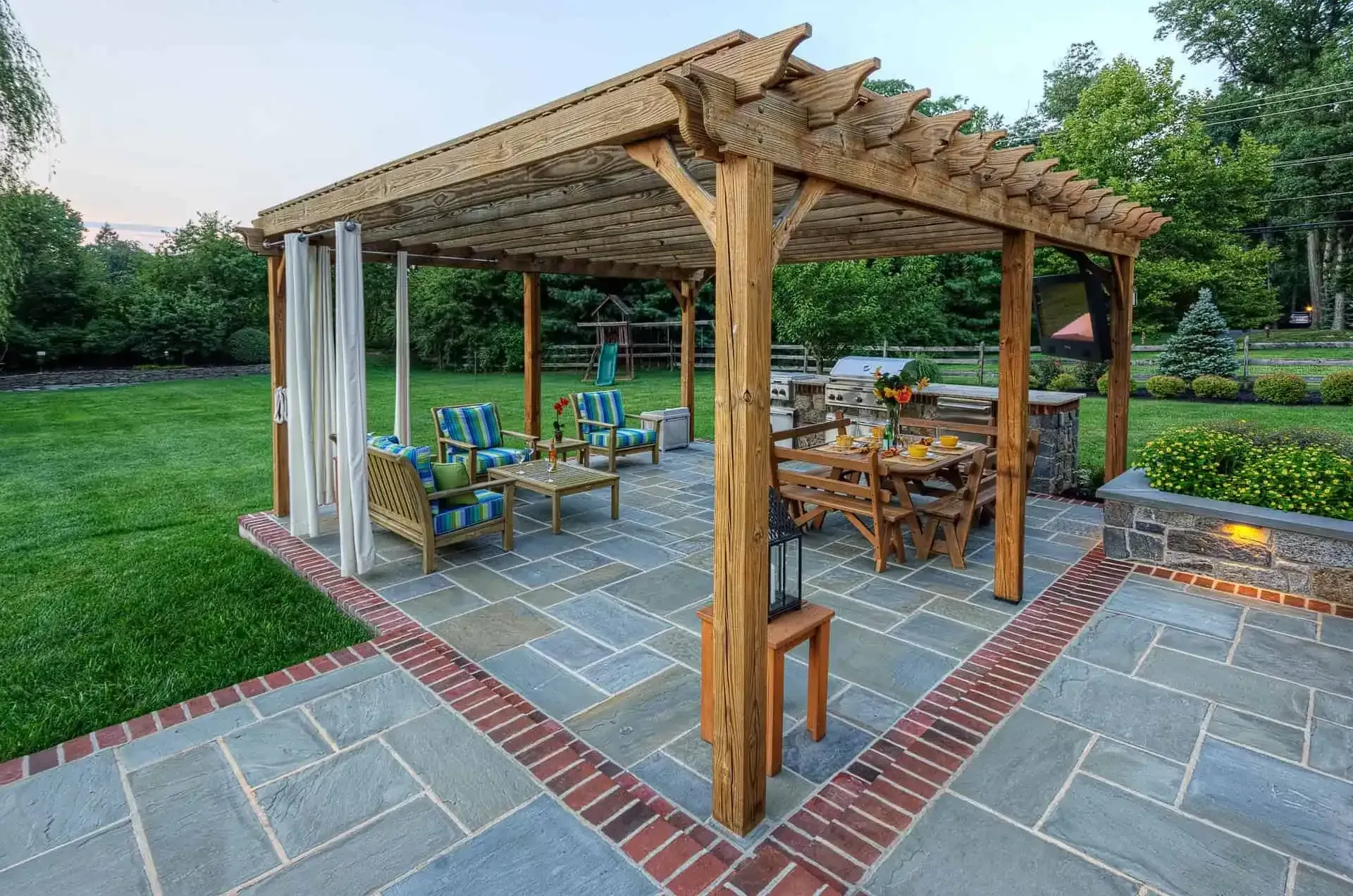 Wooden pergola with picnic tables and chairs on a patio beside a grassy yard and trees