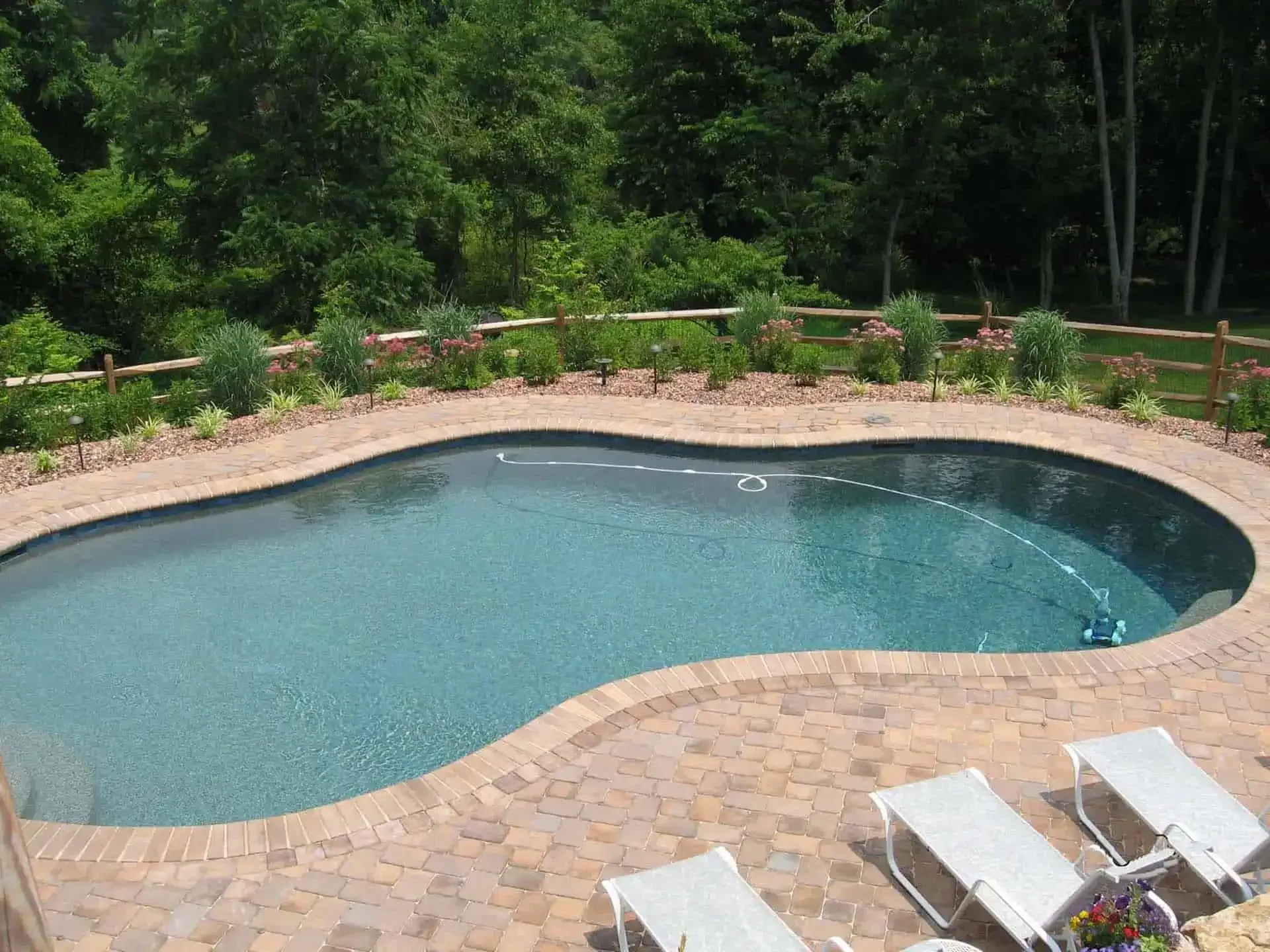 Curved backyard swimming pool with stone deck, lounge chairs, and lush greenery behind it