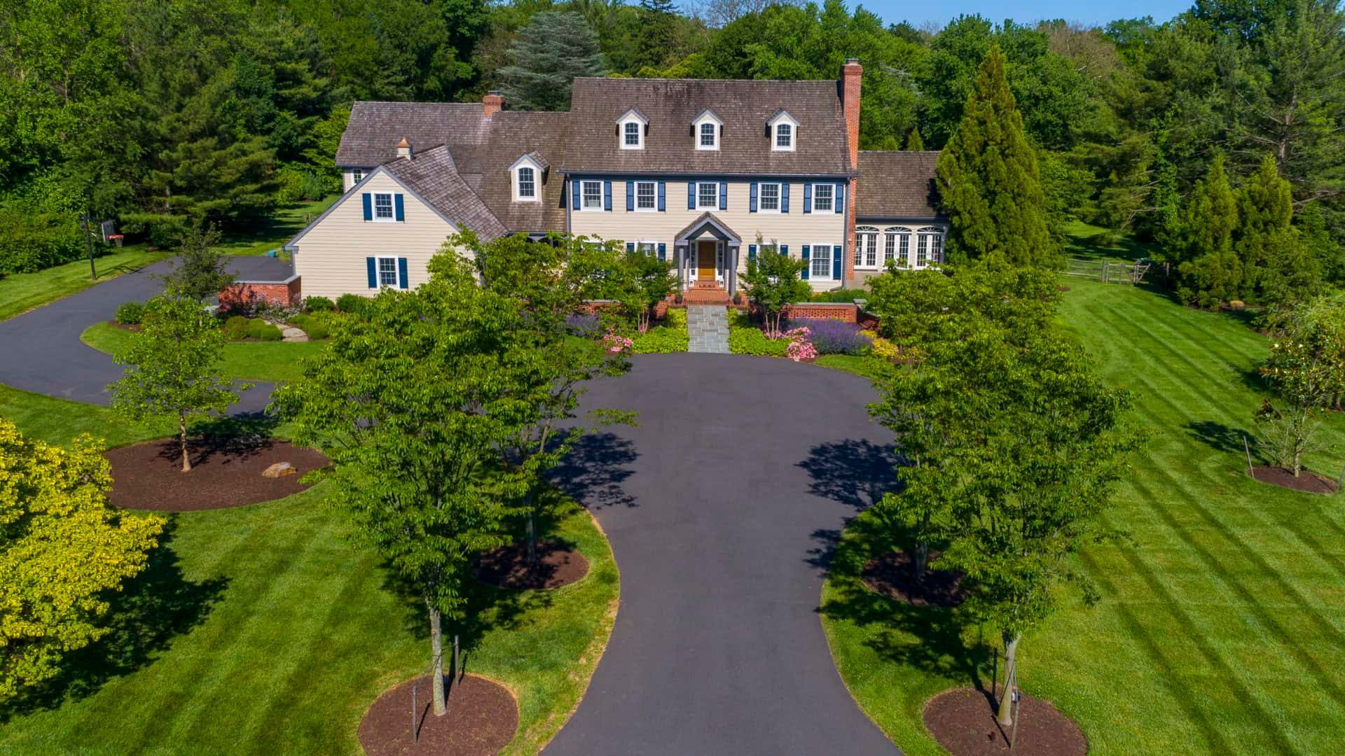 Large beige house with a dark roof, front porch, and sweeping driveway surrounded by trees and lawn.