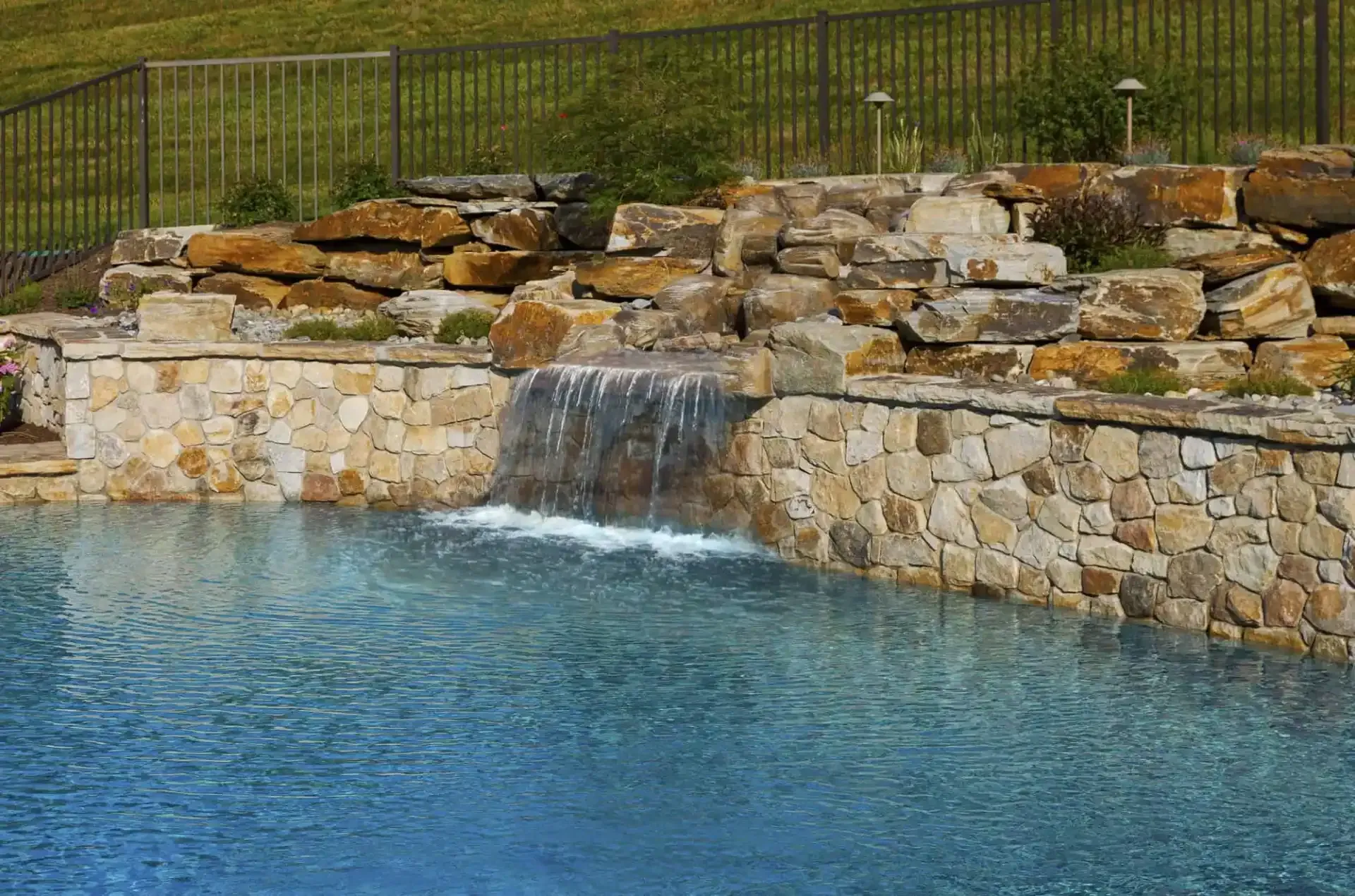 Stone waterfall spilling into a blue pool beside a stacked stone retaining wall and grass above