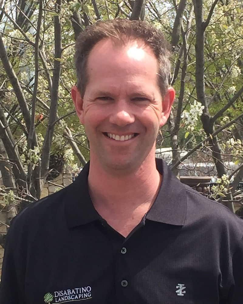 Smiling man in a black polo shirt standing outdoors with trees in the background