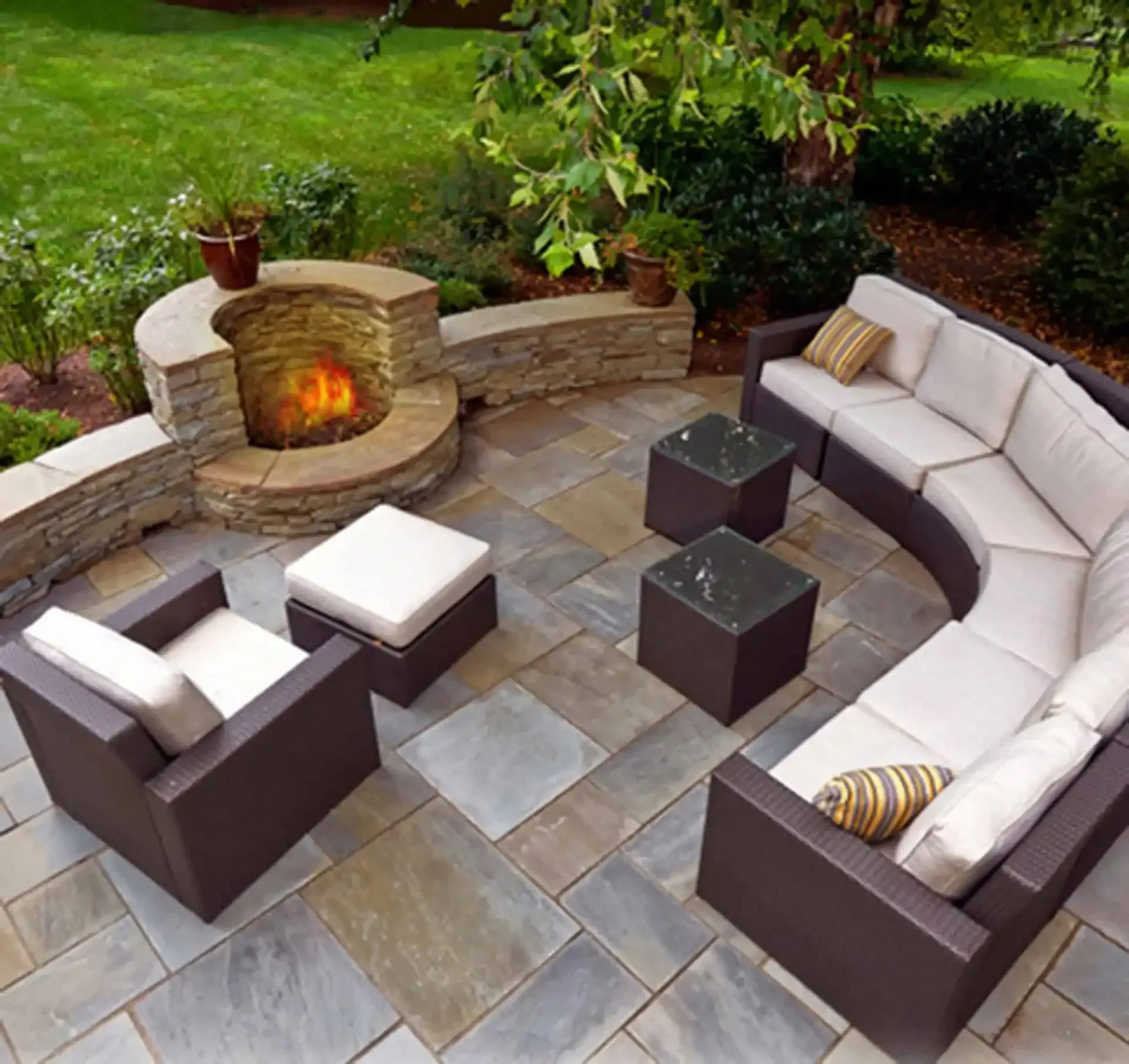 Outdoor stone patio with curved sectional seating around a built-in fire pit and garden backdrop