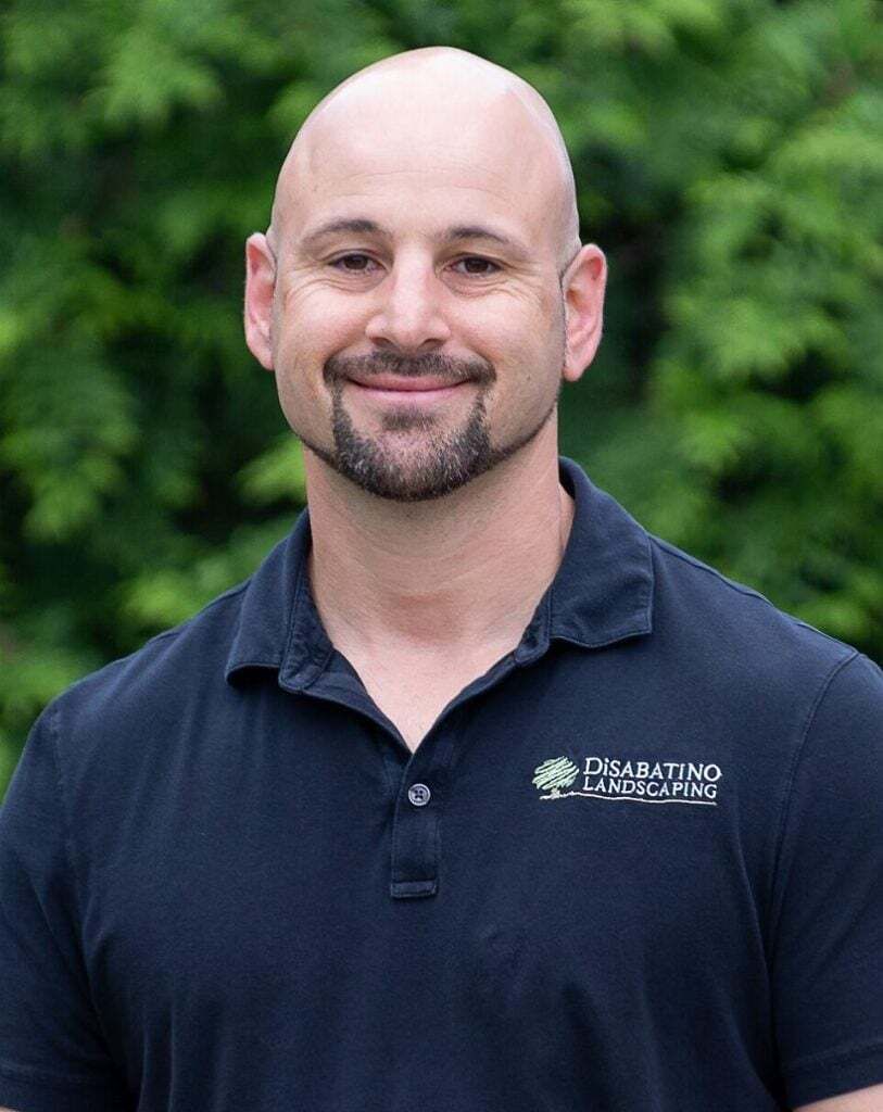 Smiling bald man in a navy polo shirt standing outdoors before green foliage