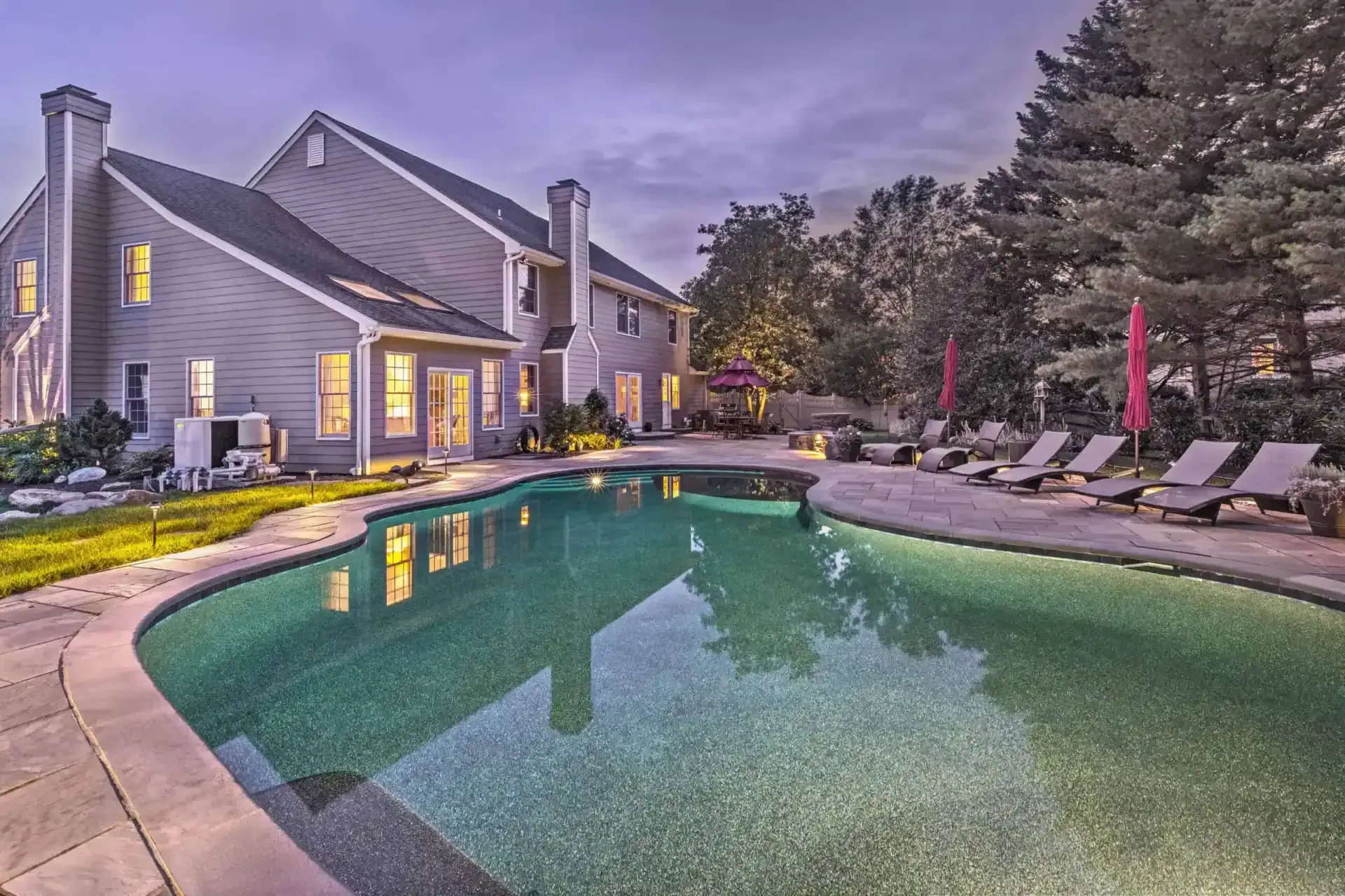 Backyard pool beside a gray house at dusk, with lounge chairs, umbrellas, and trees nearby