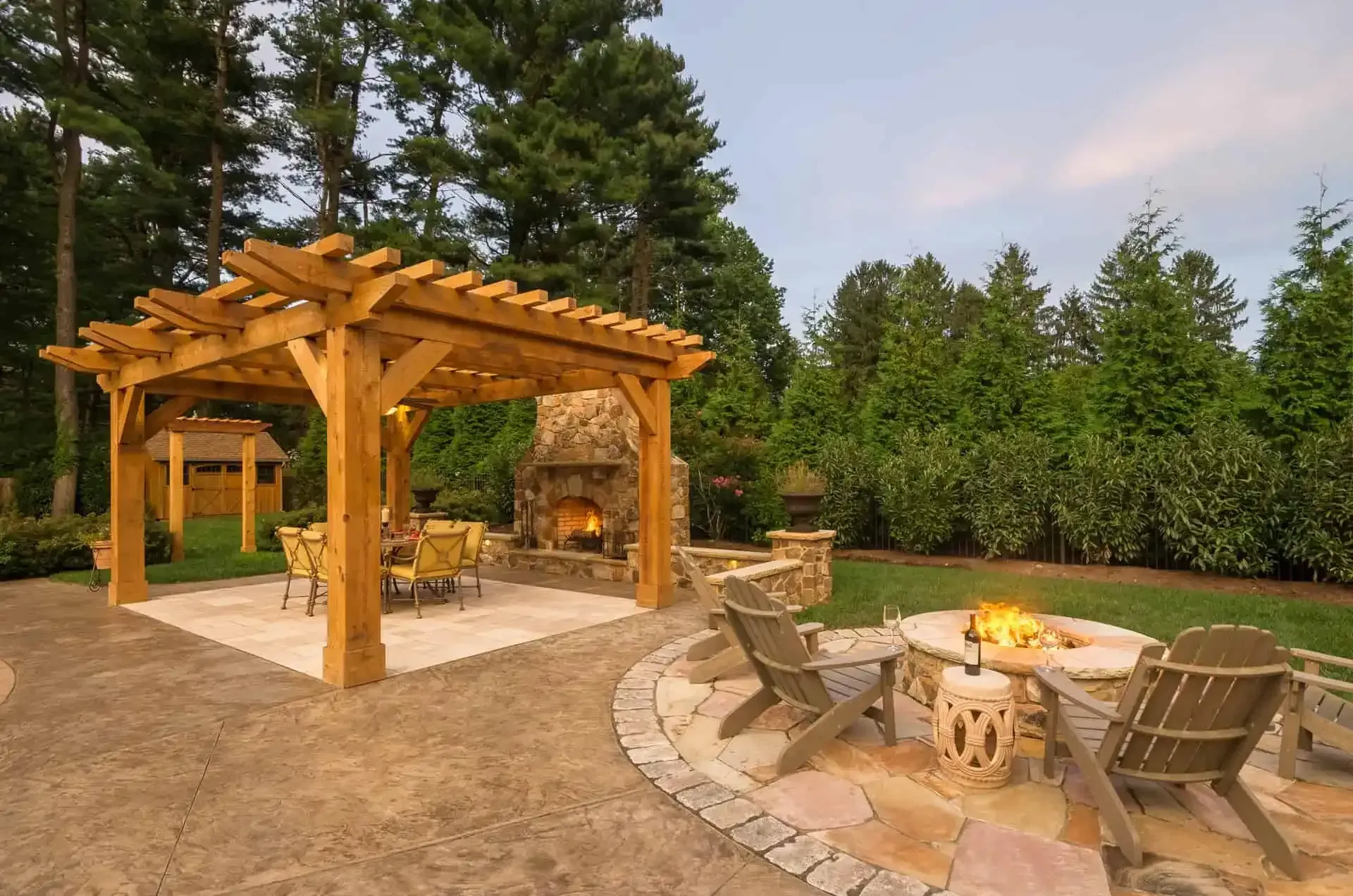 Backyard patio with wooden pergola, seating area, and stone fire pit surrounded by trees