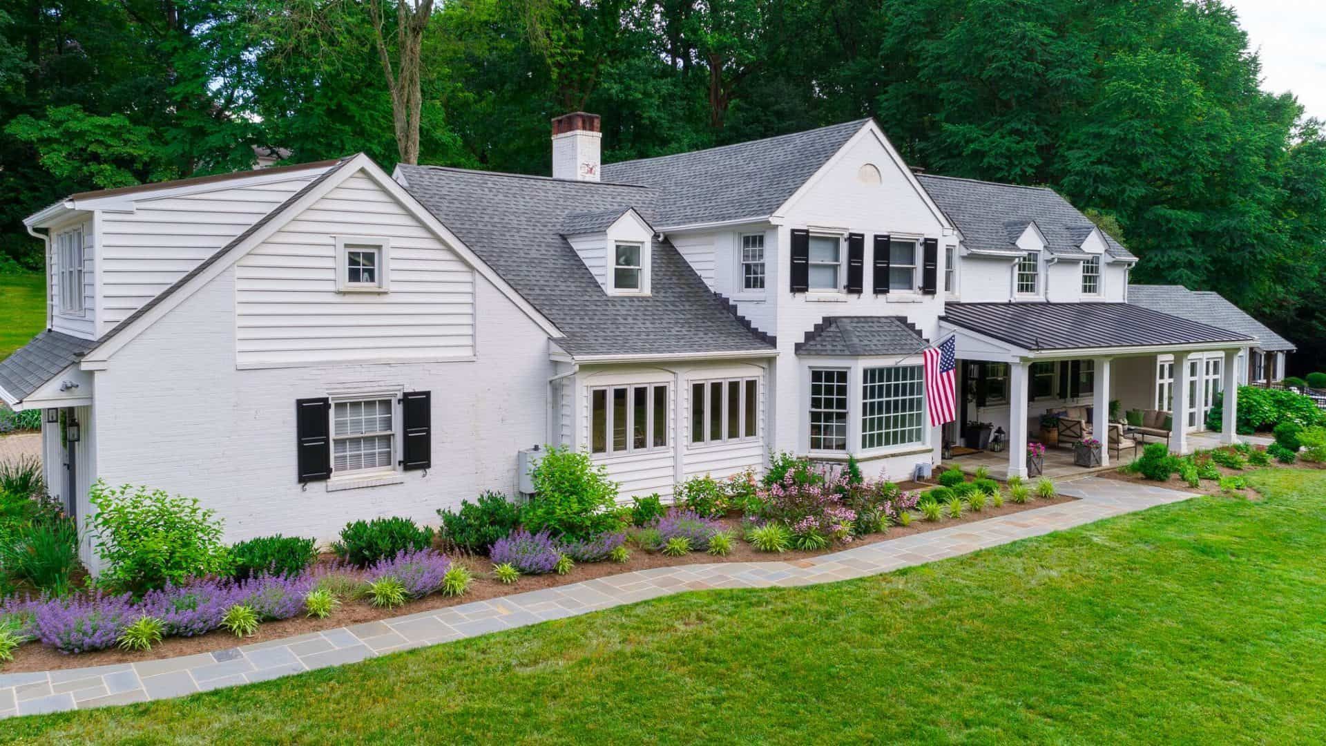 Large white house with black shutters, a porch, and a flower-lined lawn beside a wooded backdrop