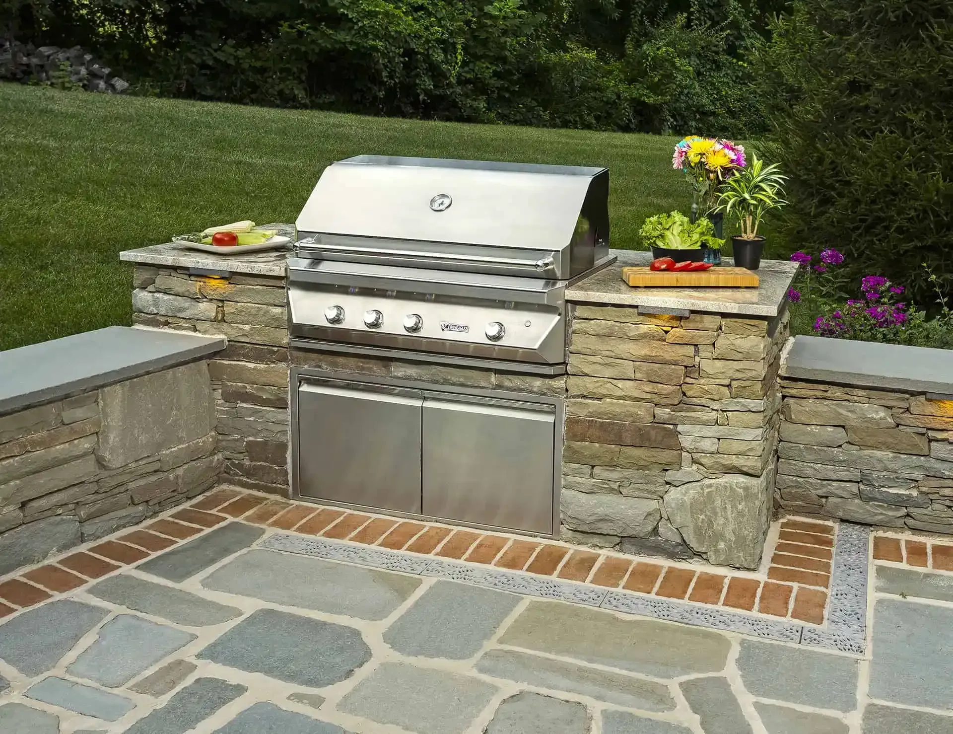 Outdoor stainless steel grill with stone surround in a landscaped backyard patio