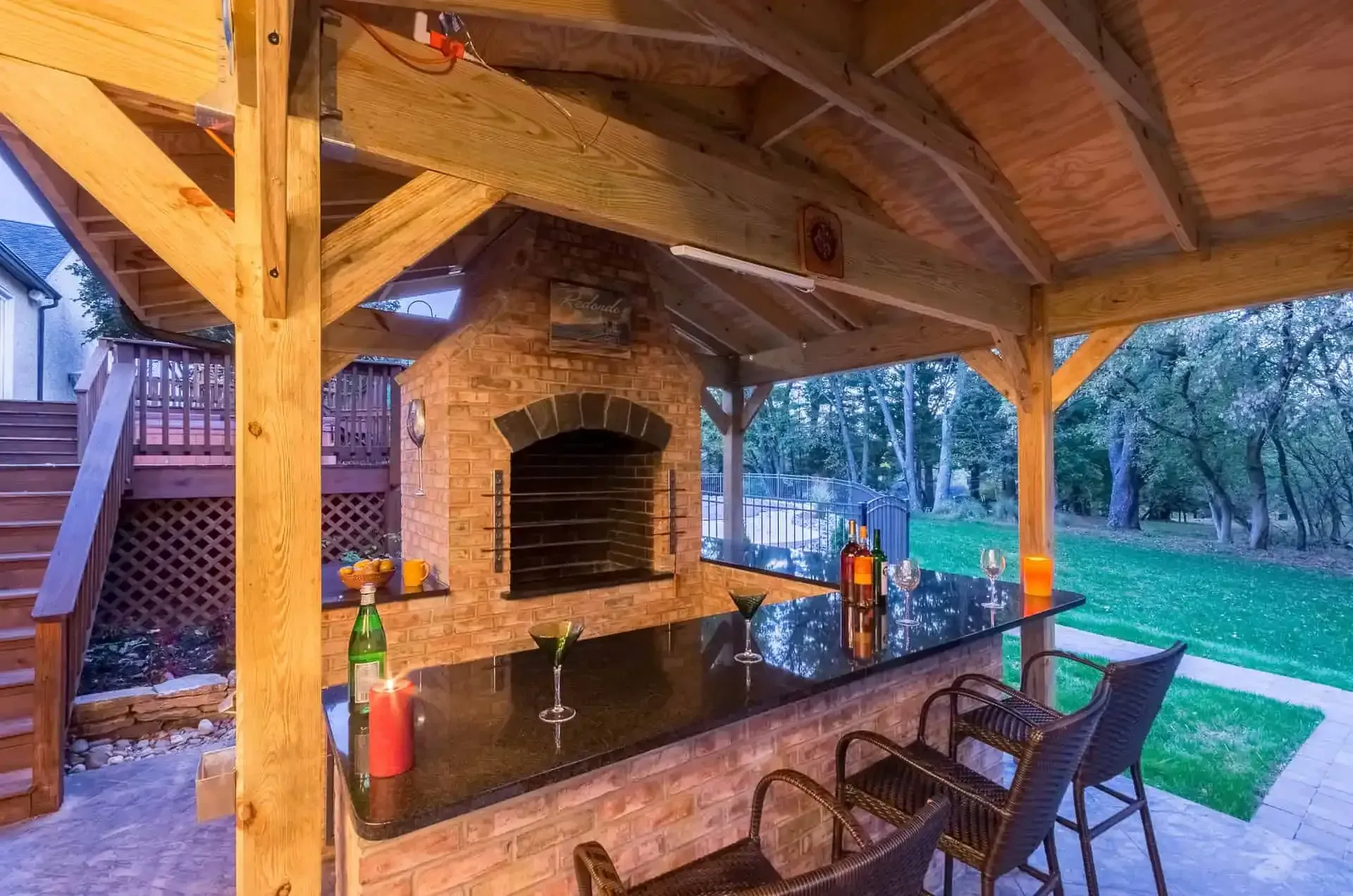 Covered outdoor patio with a brick grill, stone bar, and chairs overlooking a grassy backyard at dusk