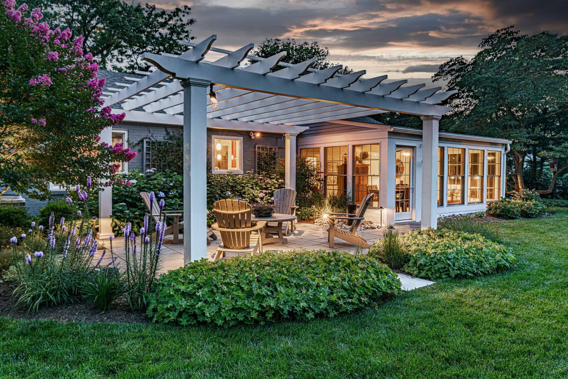 Cozy patio and garden beside a lit house at dusk, with white pergola, wicker seating, and lush greenery