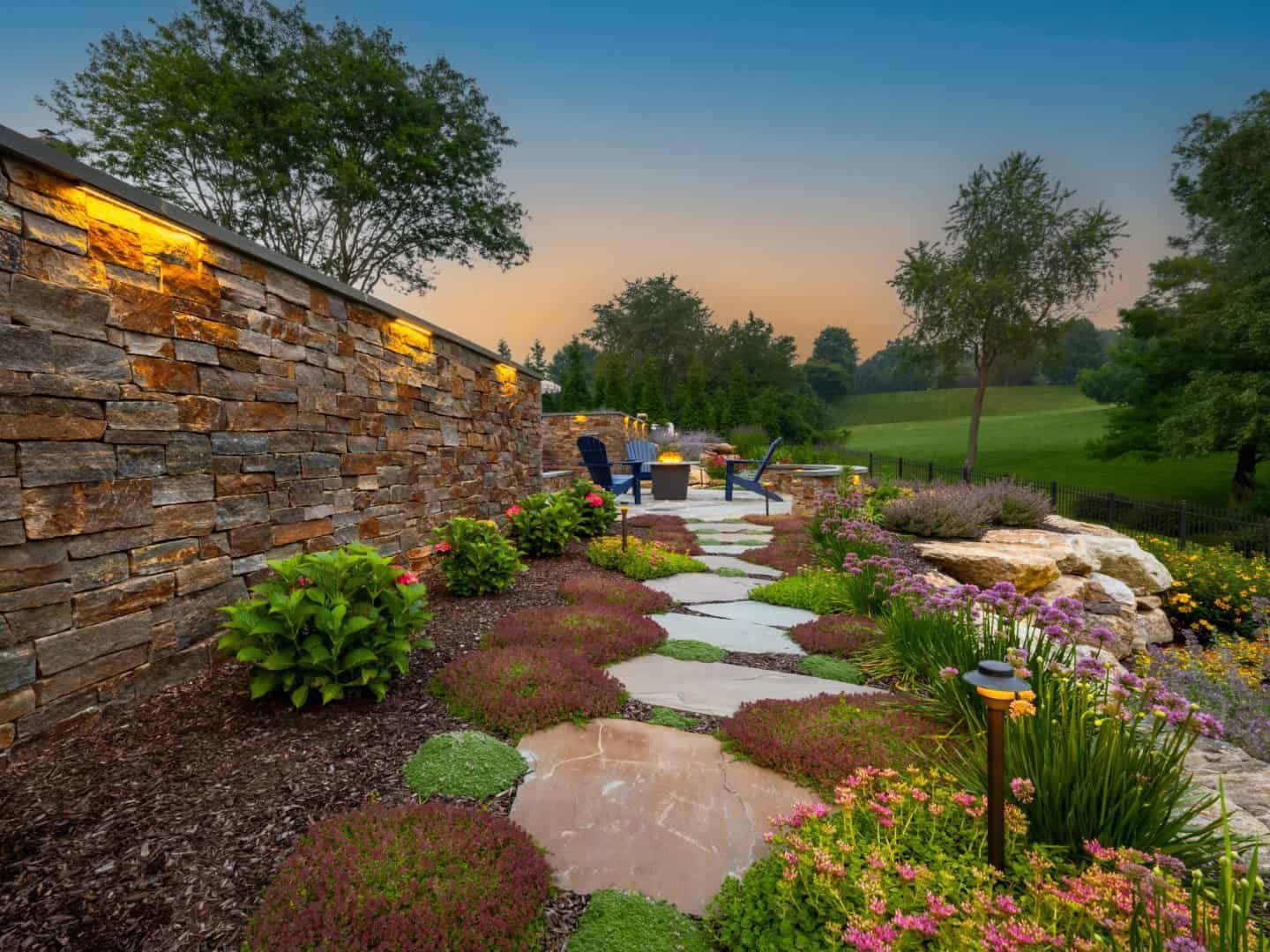 Stone garden path with stepping stones, flowers, and a lit retaining wall at sunset