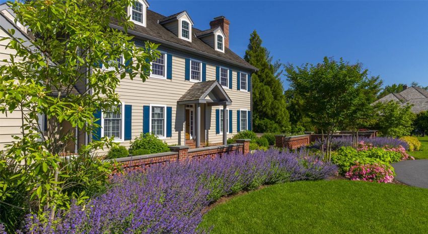 Large cream house with blue shutters and a porch, beside a purple flower border and green lawn under a sunny sky