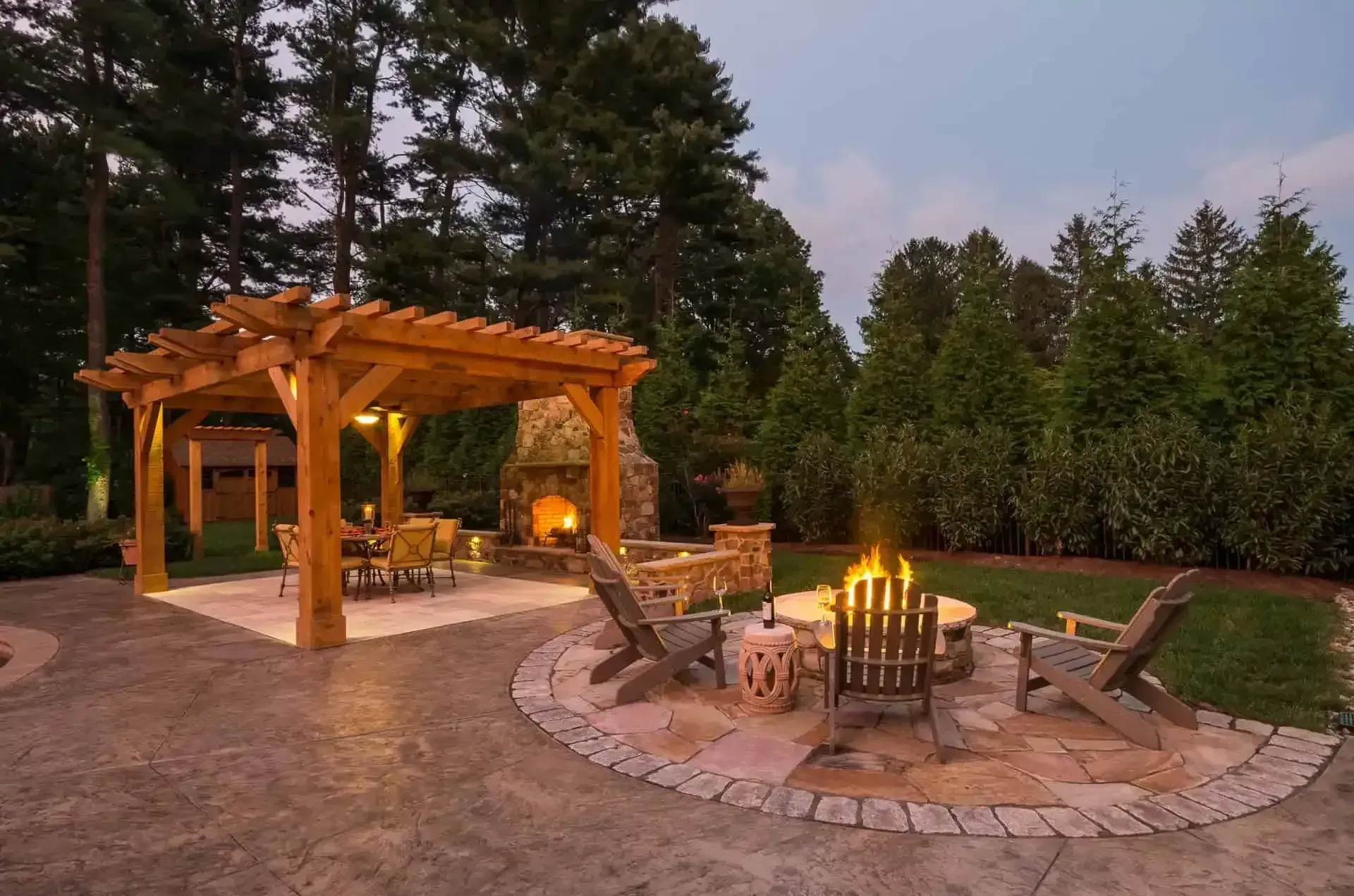 Wooden pergola and stone fire pit seating area in a wooded backyard at dusk