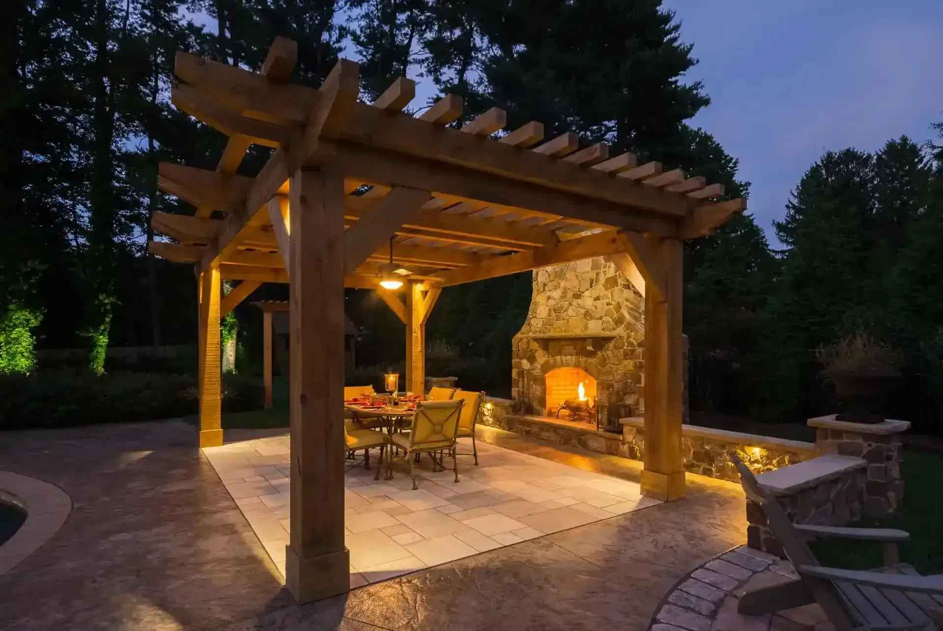 Lit outdoor patio with wooden pergola, stone fireplace, and dining table at dusk