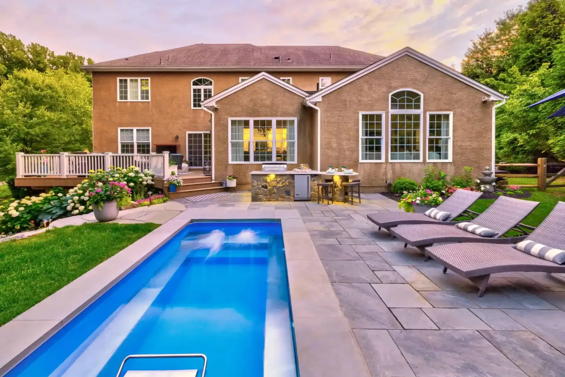 Backyard patio with a narrow pool, lounge chairs, and a brick house at sunset