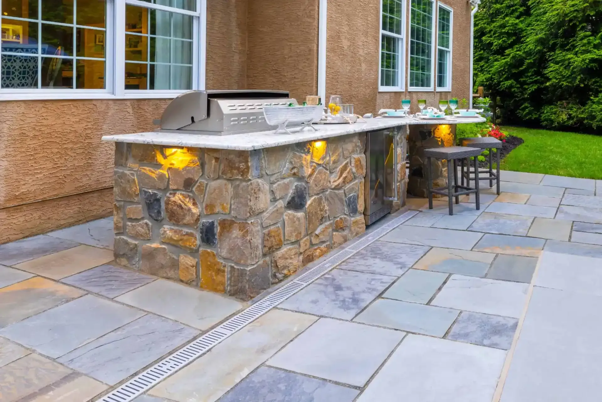 Stone outdoor kitchen with built-in grill and bar stools on a paved patio beside a house