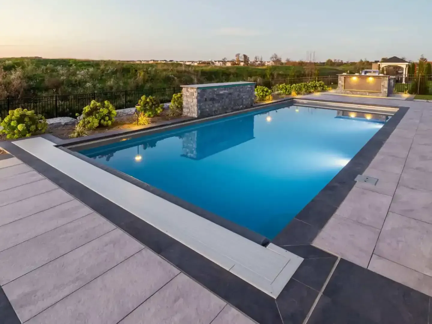 Modern backyard with a blue swimming pool, tiled deck, and landscaped greenery at sunset