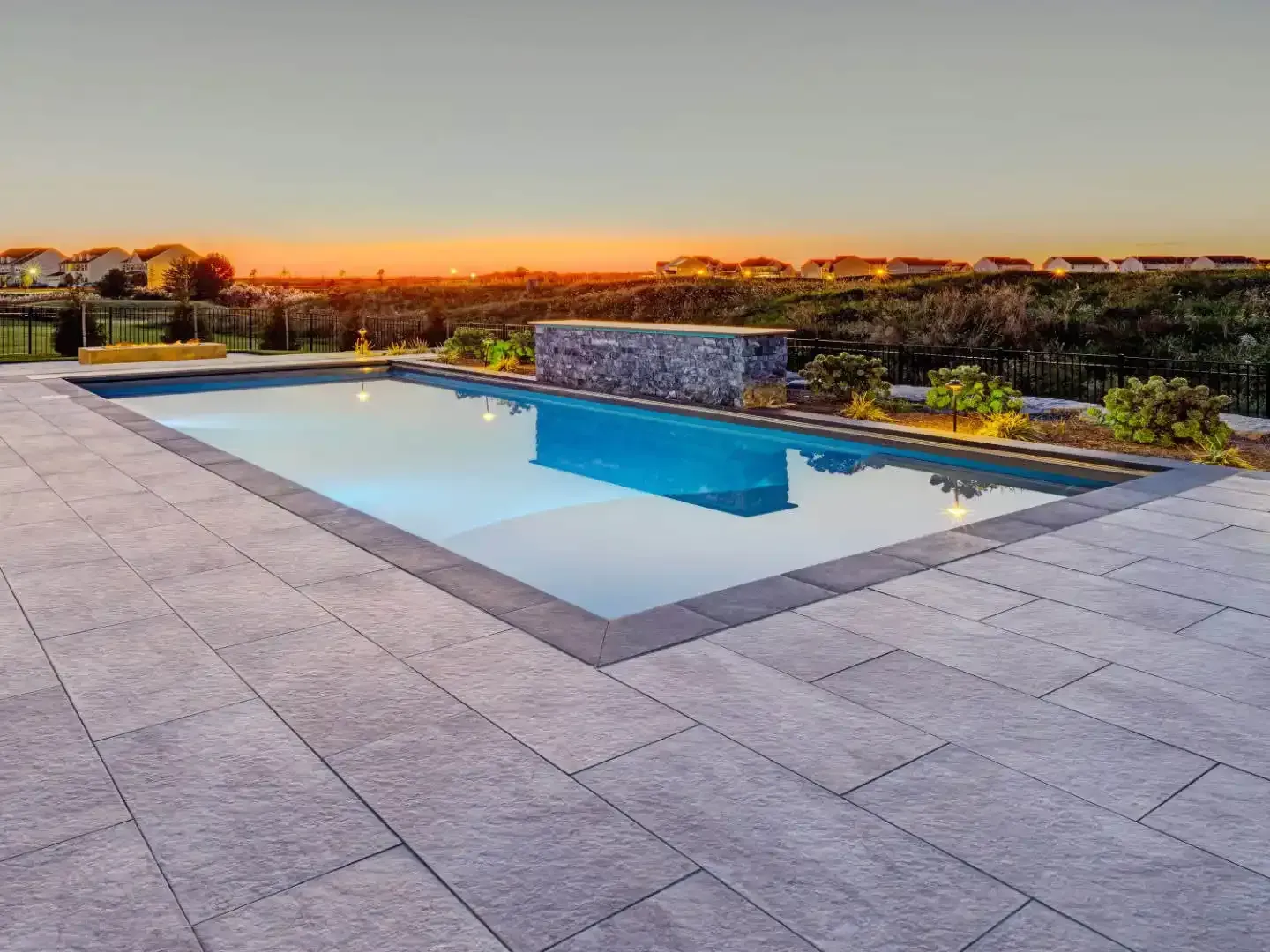 Modern backyard pool at sunset with stone patio and landscaped horizon in warm orange light