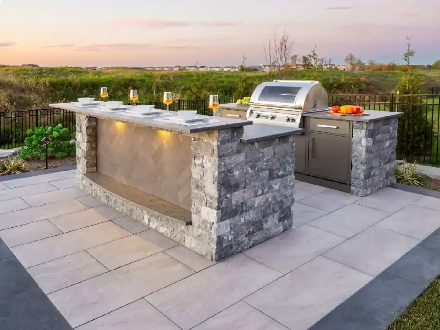 Outdoor stone kitchen island with grill, sink, and bar seating on a patio at sunset
