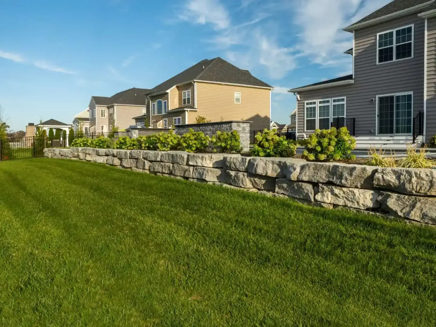 Suburban homes beside a stone retaining wall and green lawn under a clear blue sky