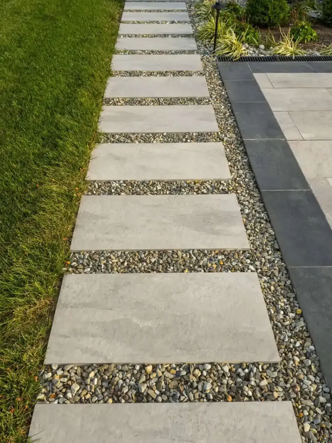 Stepping-stone walkway with concrete pavers set in gravel beside a grassy lawn and paved patio.