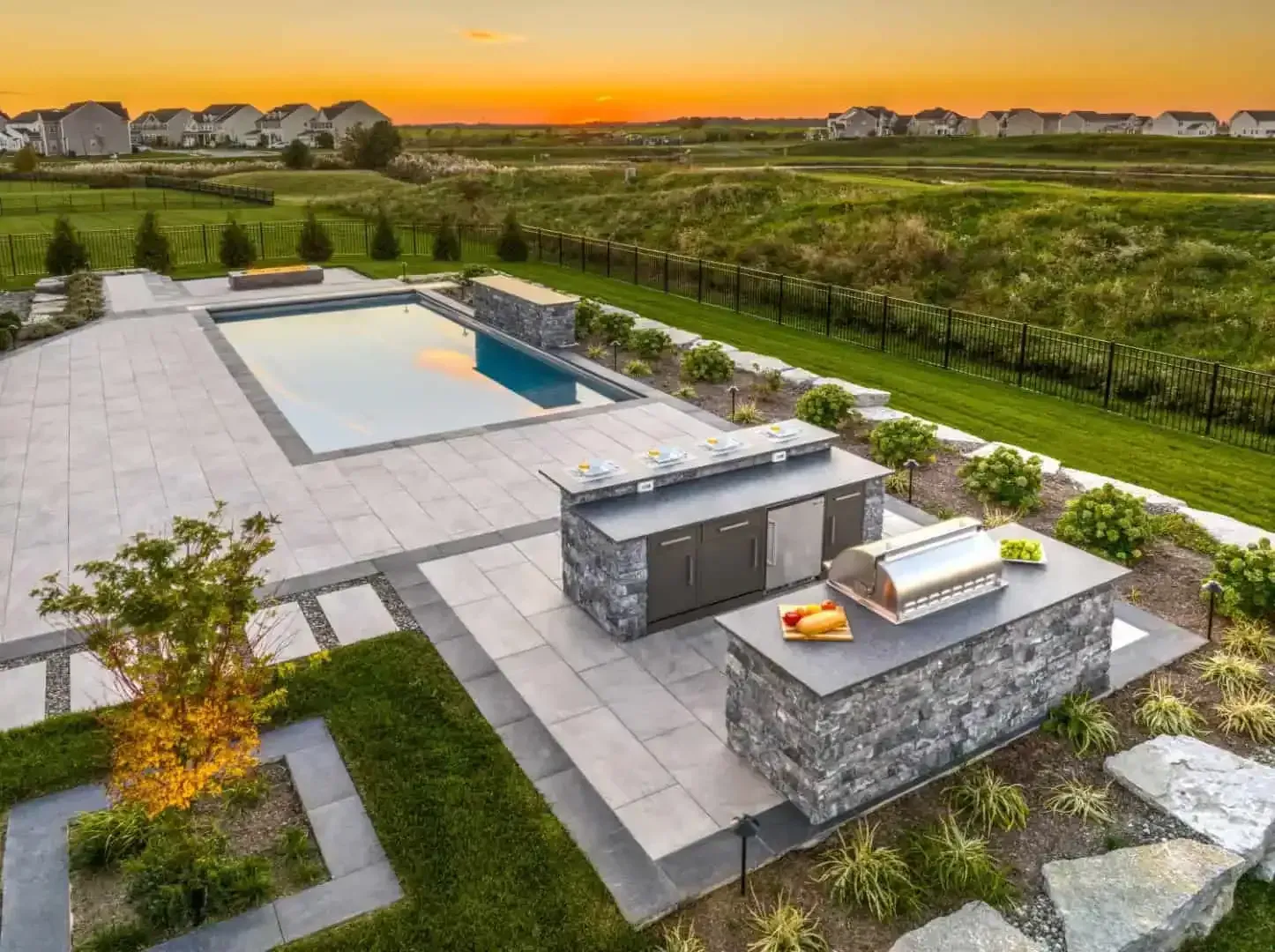 Modern backyard with a pool, stone patio, outdoor kitchen, and sunset over green fields