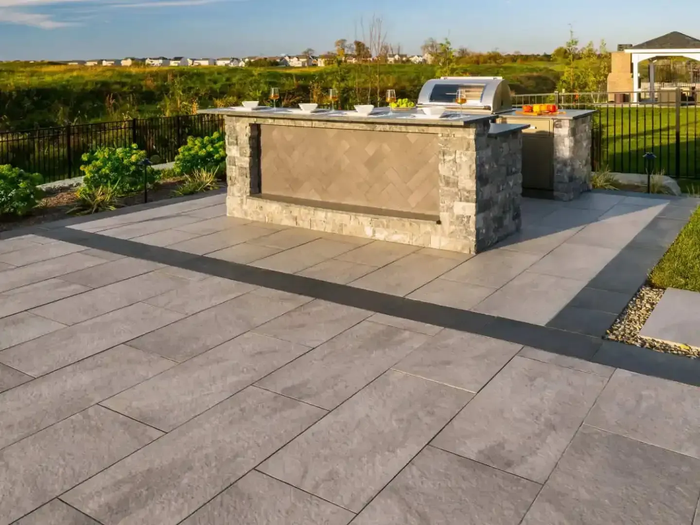 Stone outdoor kitchen island on a paved patio with a grill, set beside green fields at sunset