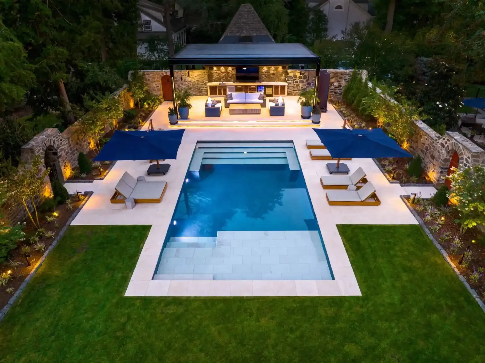 Luxury backyard pool at dusk with lounge chairs, blue umbrellas, and a lit pavilion surrounded by greenery