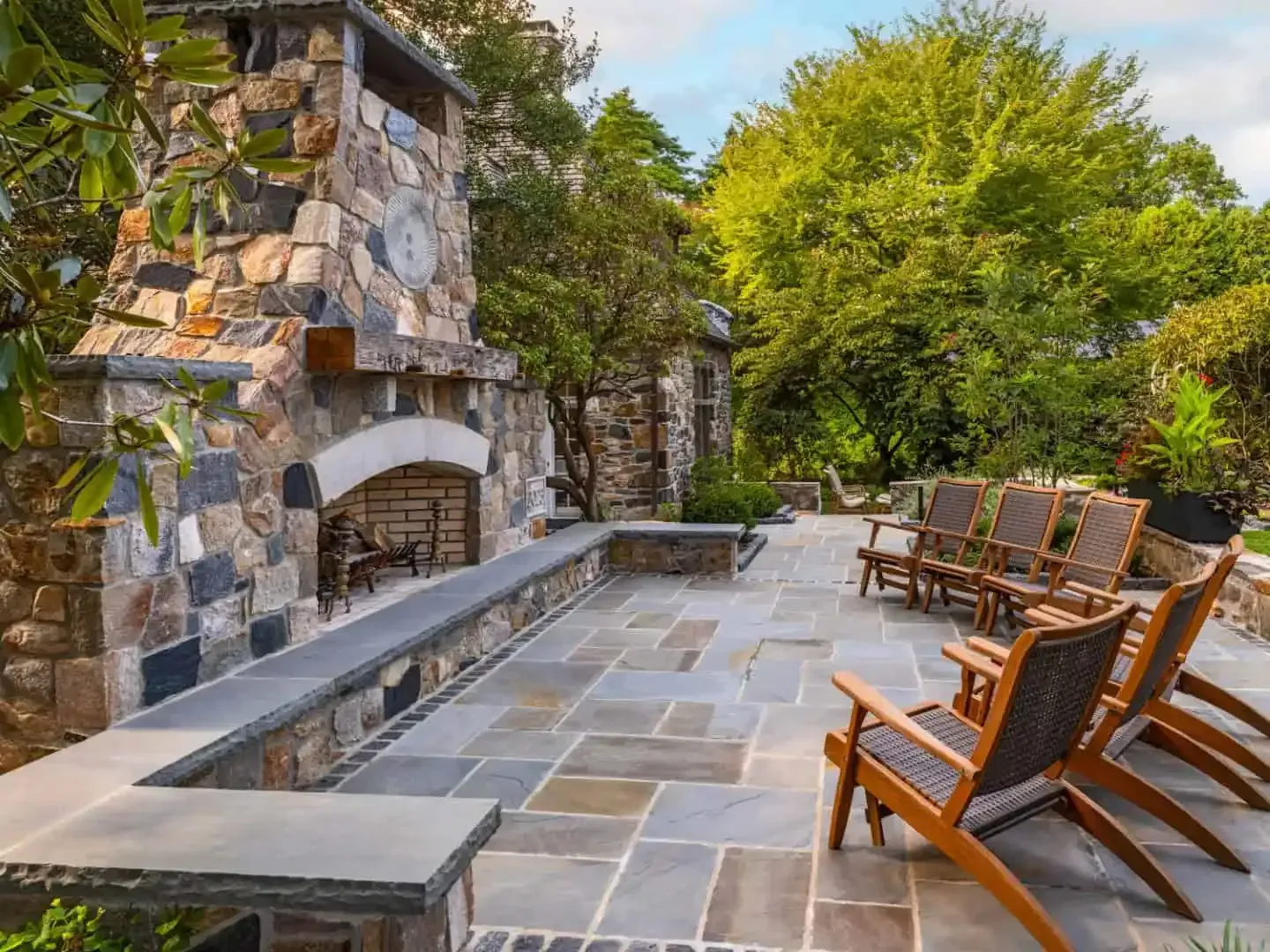Stone patio with a fireplace, gray pavers, and wooden lounge chairs beside lush greenery