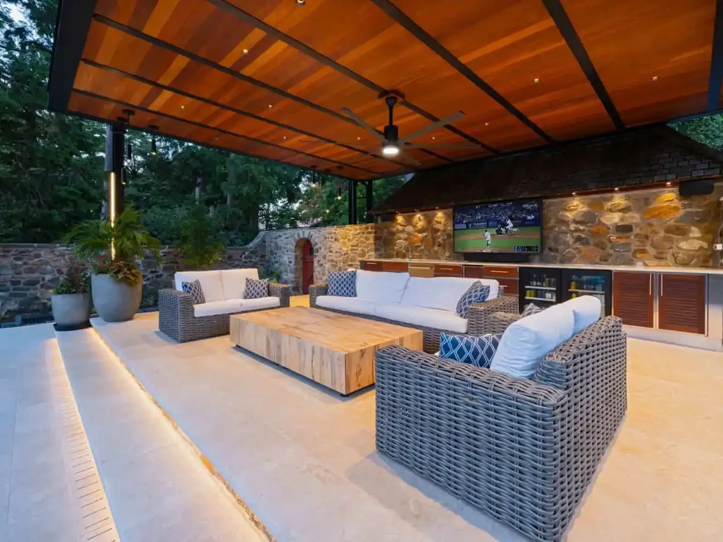 Covered outdoor patio with wicker seating, warm lighting, and stone wall at dusk