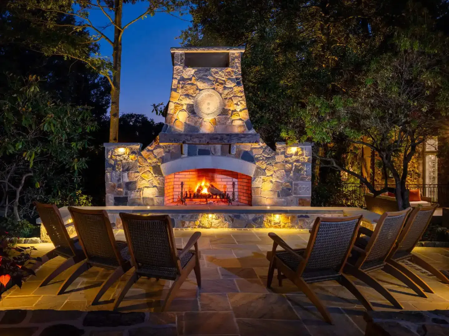 Stone outdoor fireplace glowing at night, surrounded by patio chairs and trees under warm ambient lighting