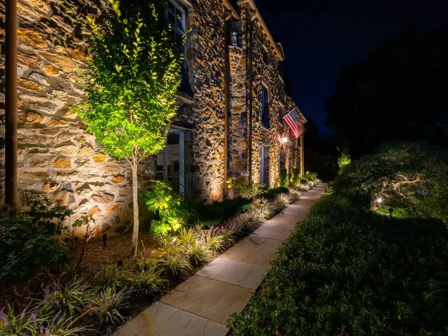 Night-lit stone building with a narrow garden walkway and illuminated trees