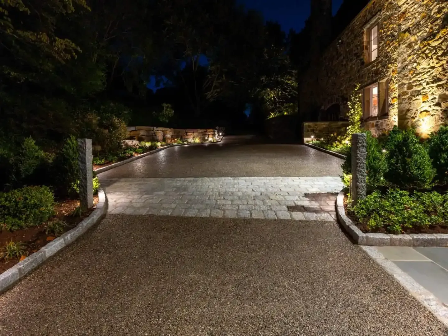 Well-lit driveway at night leading to a house with stone landscaping and gardens