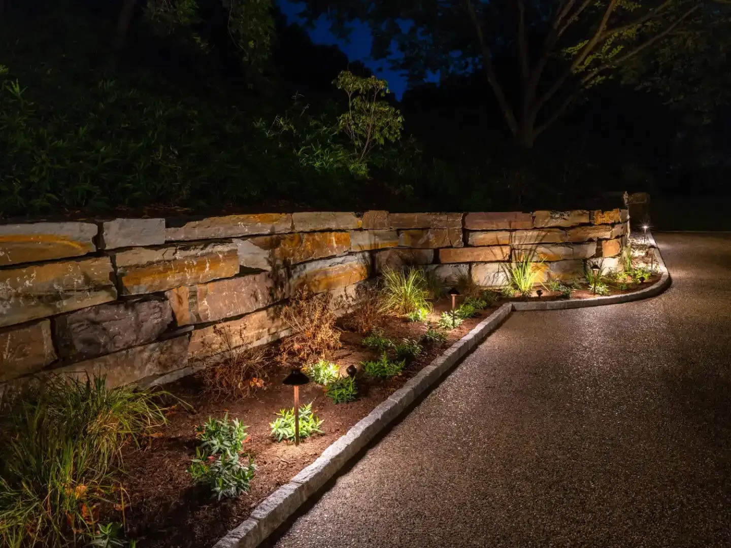 Lit stone retaining wall beside a paved path, with small plants and warm garden lights at night